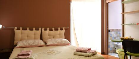 A neatly made bed with pillows and folded towels, a bedside table with a lamp, and a chair near a window with curtains.