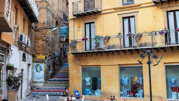 A narrow street with outdoor seating, a yellow building with balconies, and a staircase leading to an upper floor. - Le Casette di Lù (Agrigento)