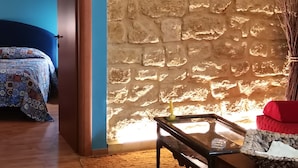 A room with a stone wall, a wooden floor, a bed with patterned bedding, a glass-top table, and a red cushion. - Le Casette di Lù (Agrigento)