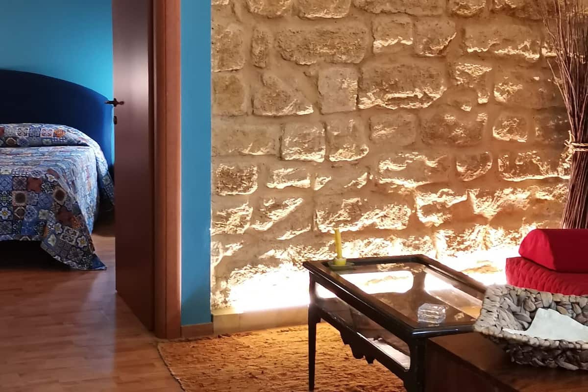 Ein Zimmer mit Steinwand, Holzboden, einem Bett mit gemusterten Textilien, einem Tisch mit Glasplatte und einem roten Kissen.