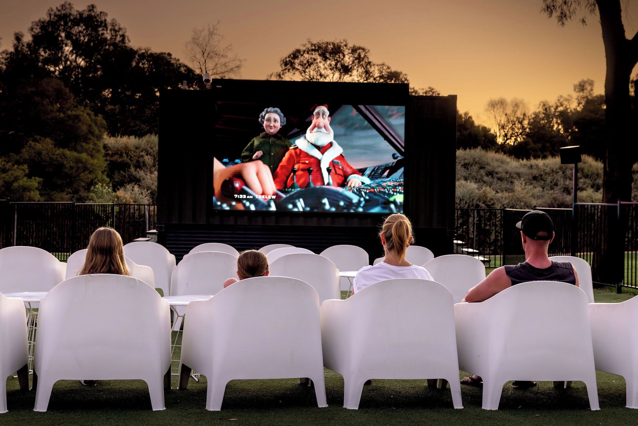 An outdoor movie screening with a large screen, white plastic chairs, and a grassy area.