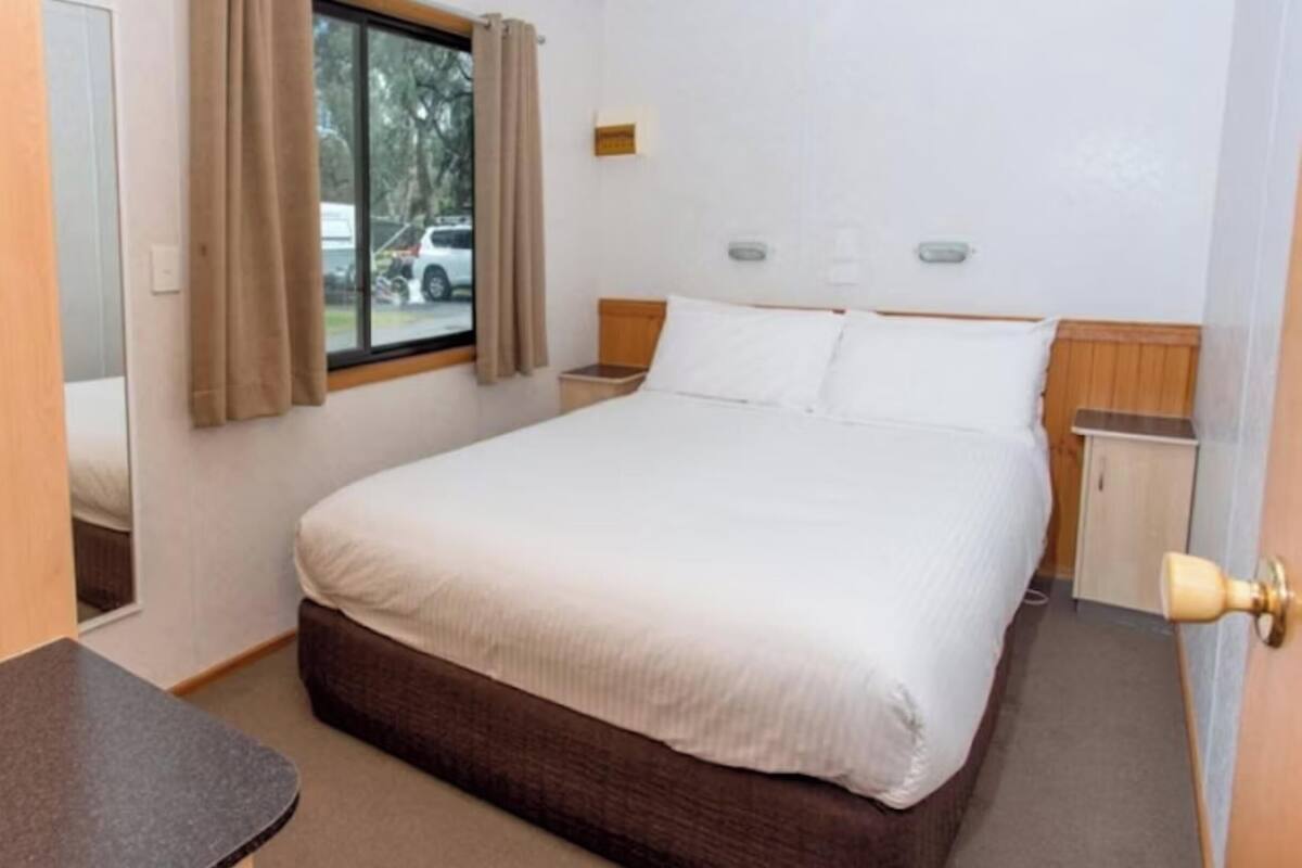 Habitación de hotel con cama, mesita de noche, ventana con cortinas, puerta y una pequeña mesa.