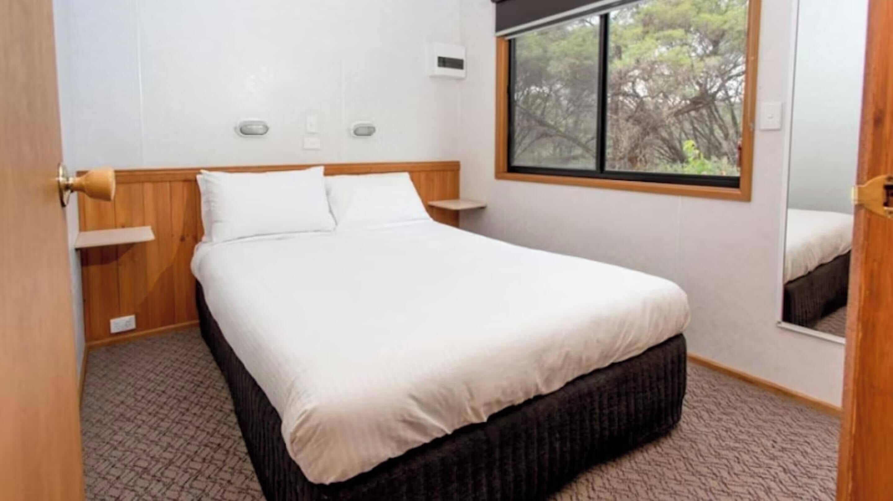 A bedroom with a bed, wooden headboard, bedside tables, a window with a view of trees, and a carpeted floor.