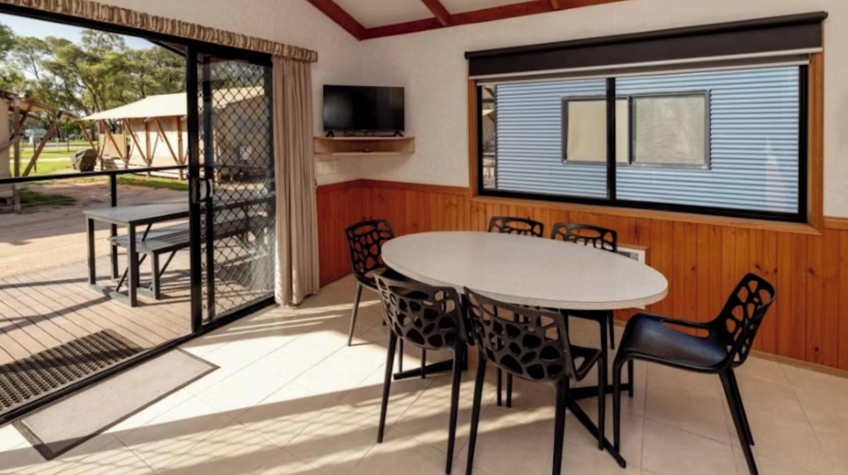 Un comedor con una mesa redonda y sillas, un televisor montado en la pared, y vistas al exterior a través de un ventanal corredera.