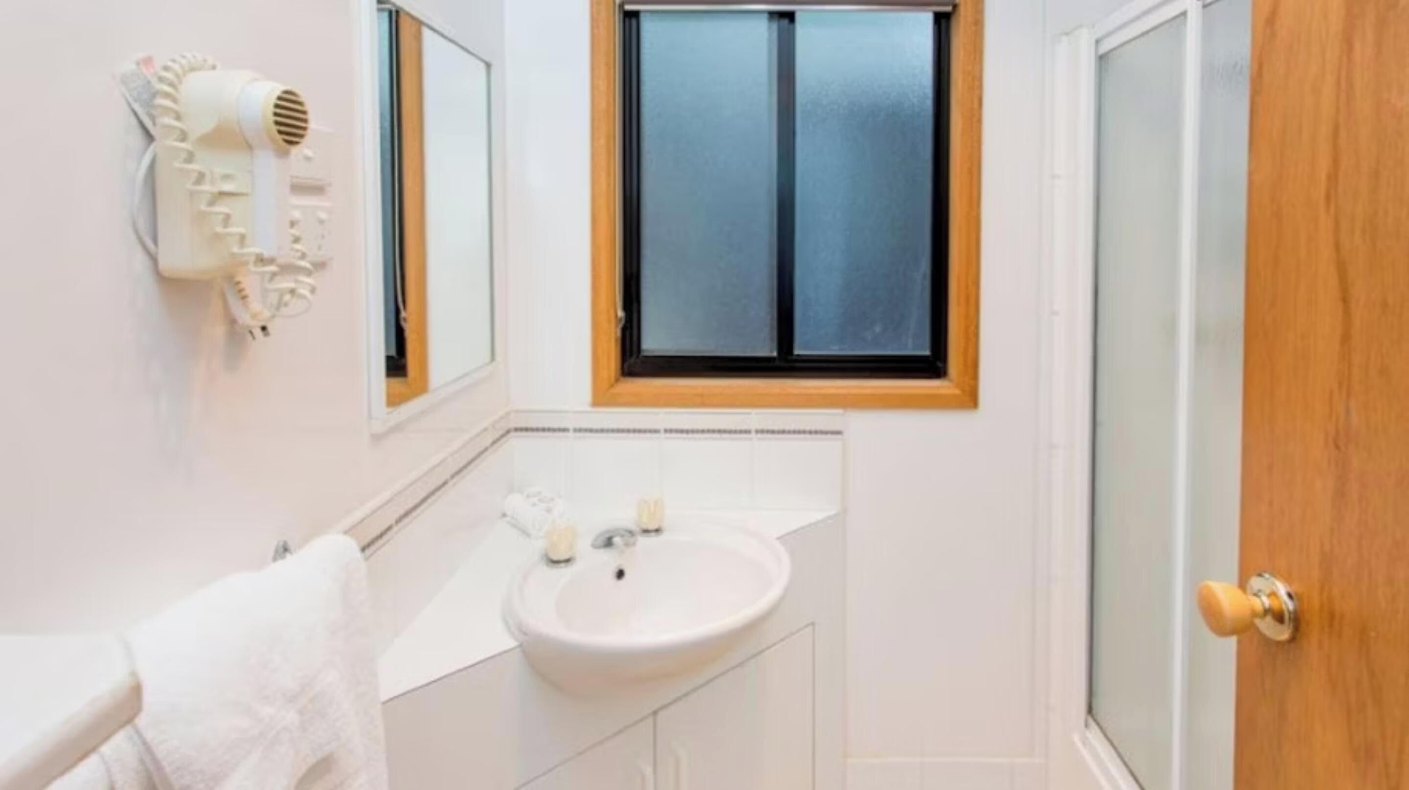 Baño con lavamanos blanco, espejo y barra para toallas. Se aprecia una ventana con marco de madera y un secador de pelo montado en la pared.