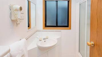 Baño con lavamanos blanco, espejo y barra para toallas. Se aprecia una ventana con marco de madera y un secador de pelo montado en la pared.