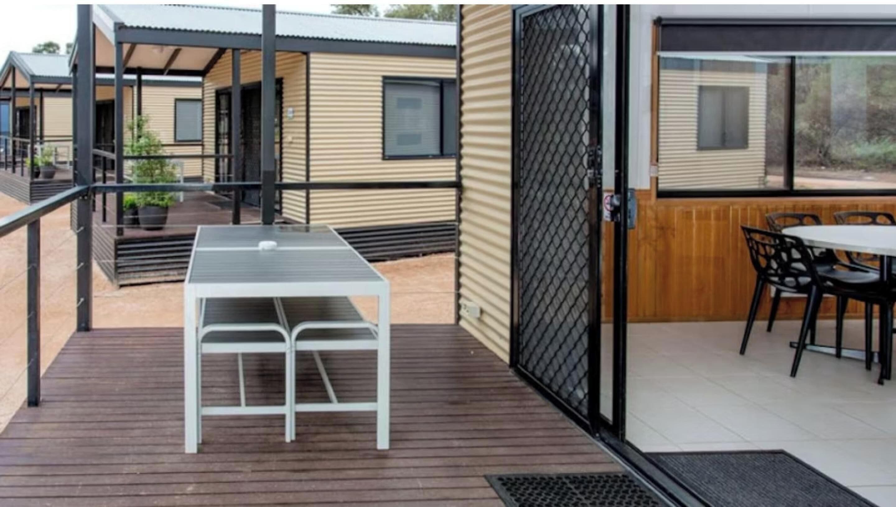 Un espace patio extérieur moderne, avec une table et des chaises, un coin salon abrité et une vue sur une maison mobile en arrière-plan.