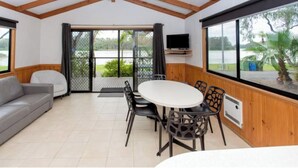A room with a grey sofa, a round table with chairs, a TV mounted on the wall, and large windows with a view of greenery outside. - Discovery Parks - Renmark Riverfront (Renmark)