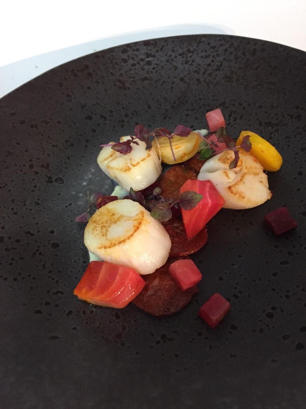A dish with scallops, red vegetables, and yellow fruit on a dark plate.