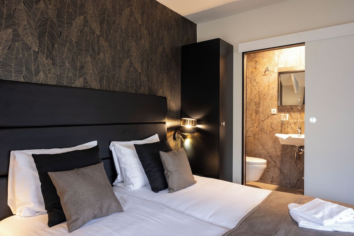 A hotel room with a large bed, a headboard, a wall-mounted light, a bathroom with a marble wall, and a sink.
