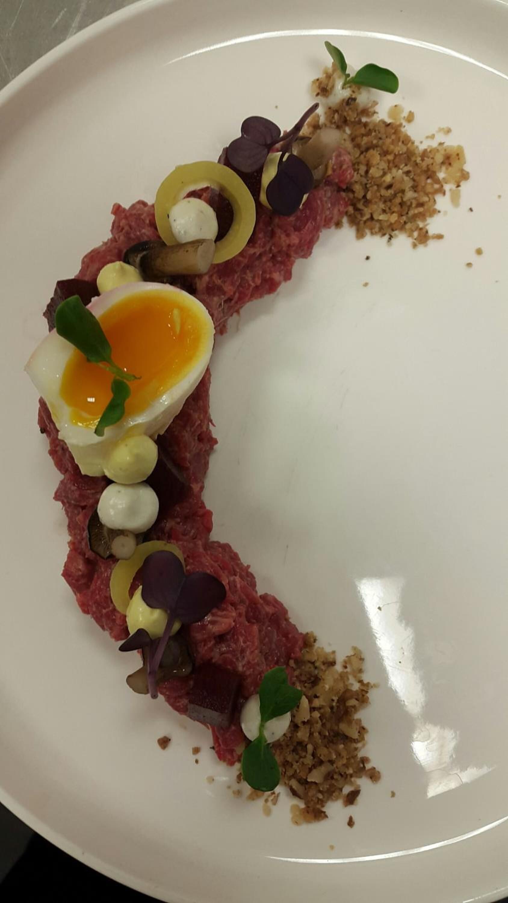 A white plate with a dish of raw meat, egg, and various garnishes.
