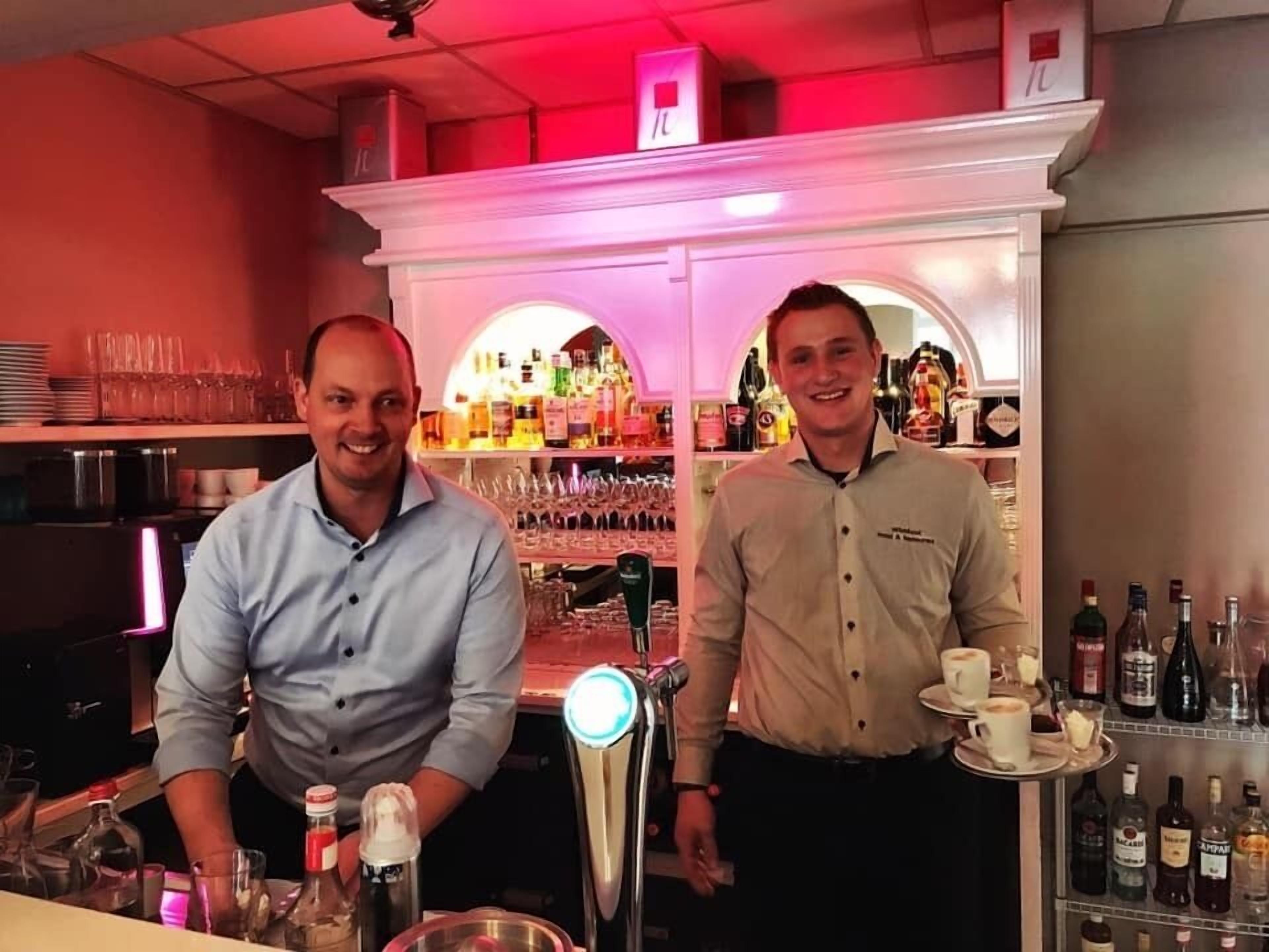 Two bartenders in a bar setting with shelves of bottles and glasses.