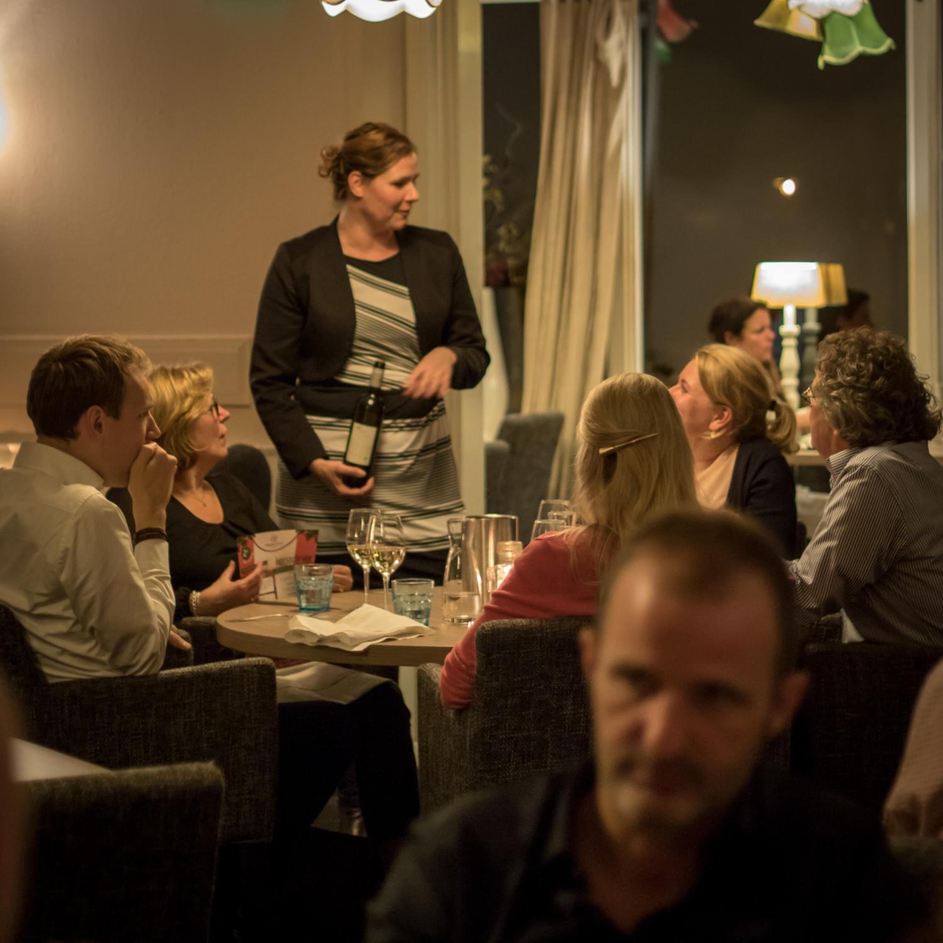 Een groep mensen zit aan tafel in een schemerige kamer met een groot raam en gordijnen.