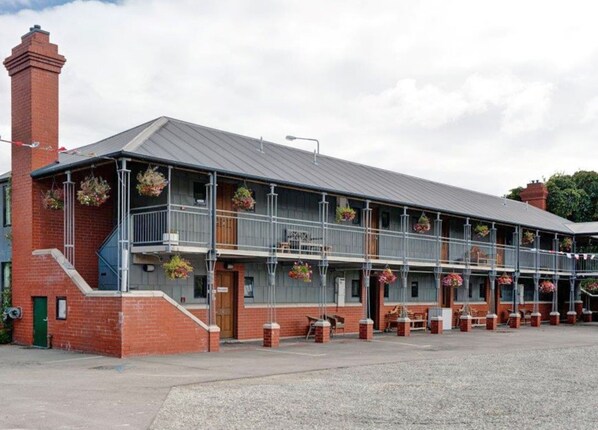 A two-story building with a grey metal roof and brick walls, featuring a balcony with hanging flower baskets.