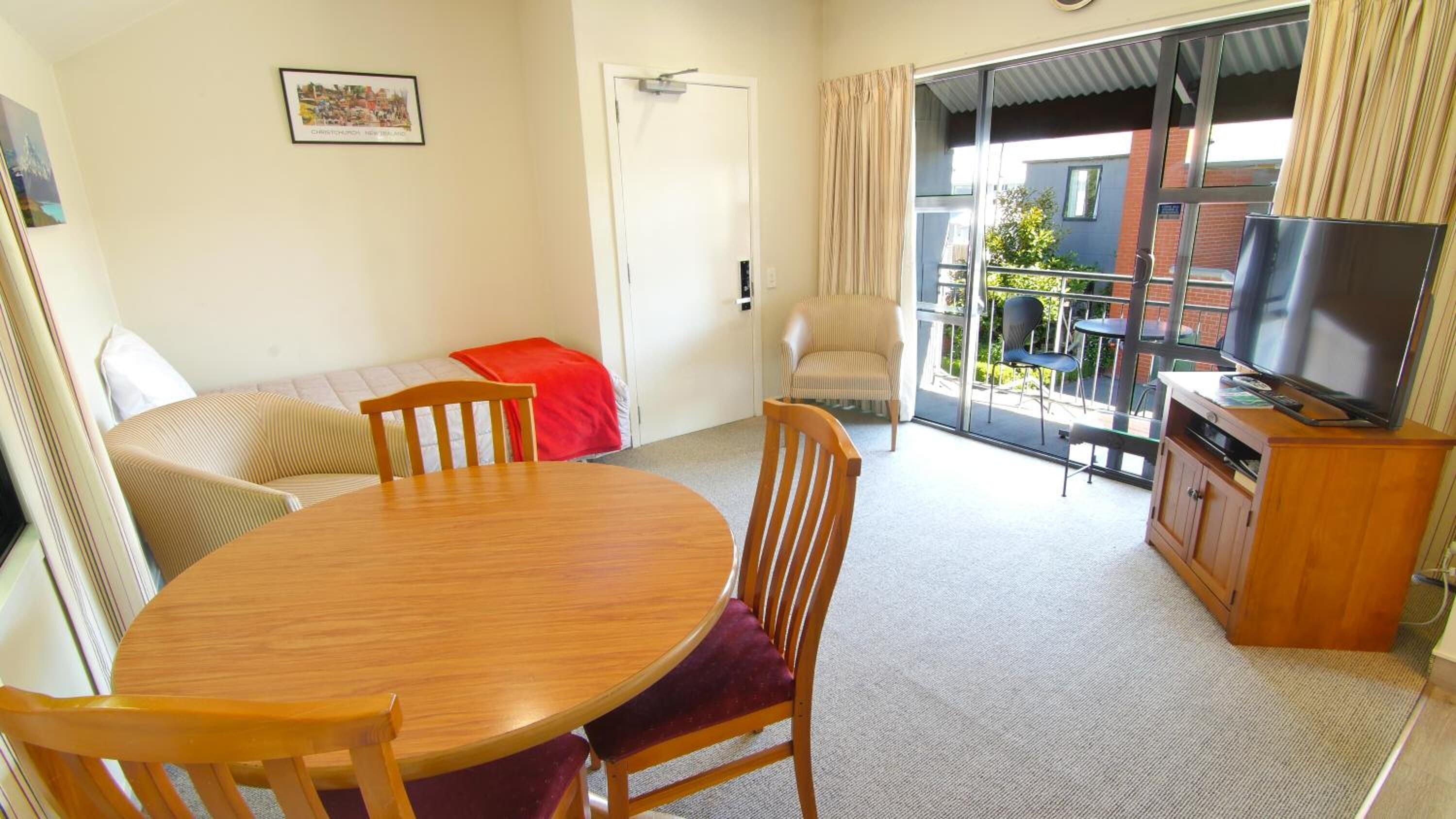 A room with a wooden dining table, chairs, a sofa, a TV on a wooden cabinet, and a sliding glass door leading to an outdoor area.