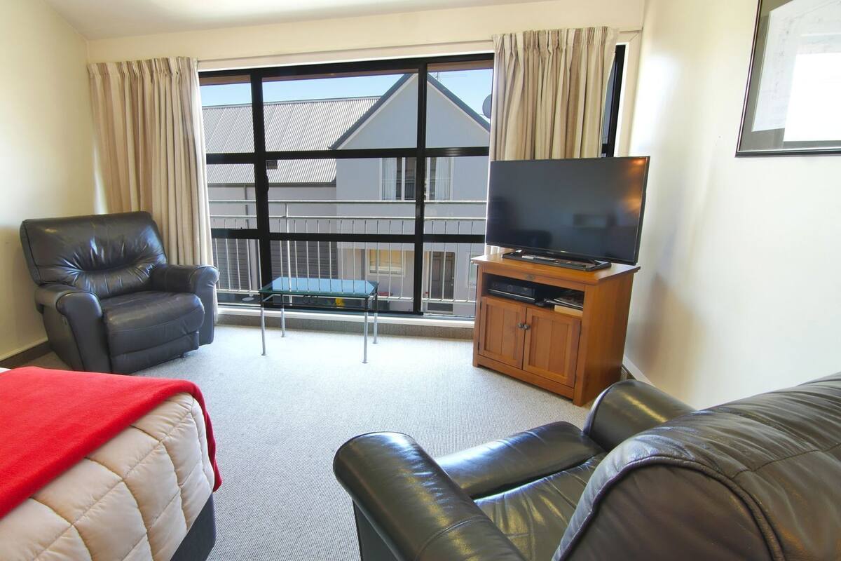 Ein Hotelzimmer mit einem Bett, einem Ledersessel, einer Couch, einem Fernseher auf einer Holzkommode und einem Fenster mit Vorhängen.