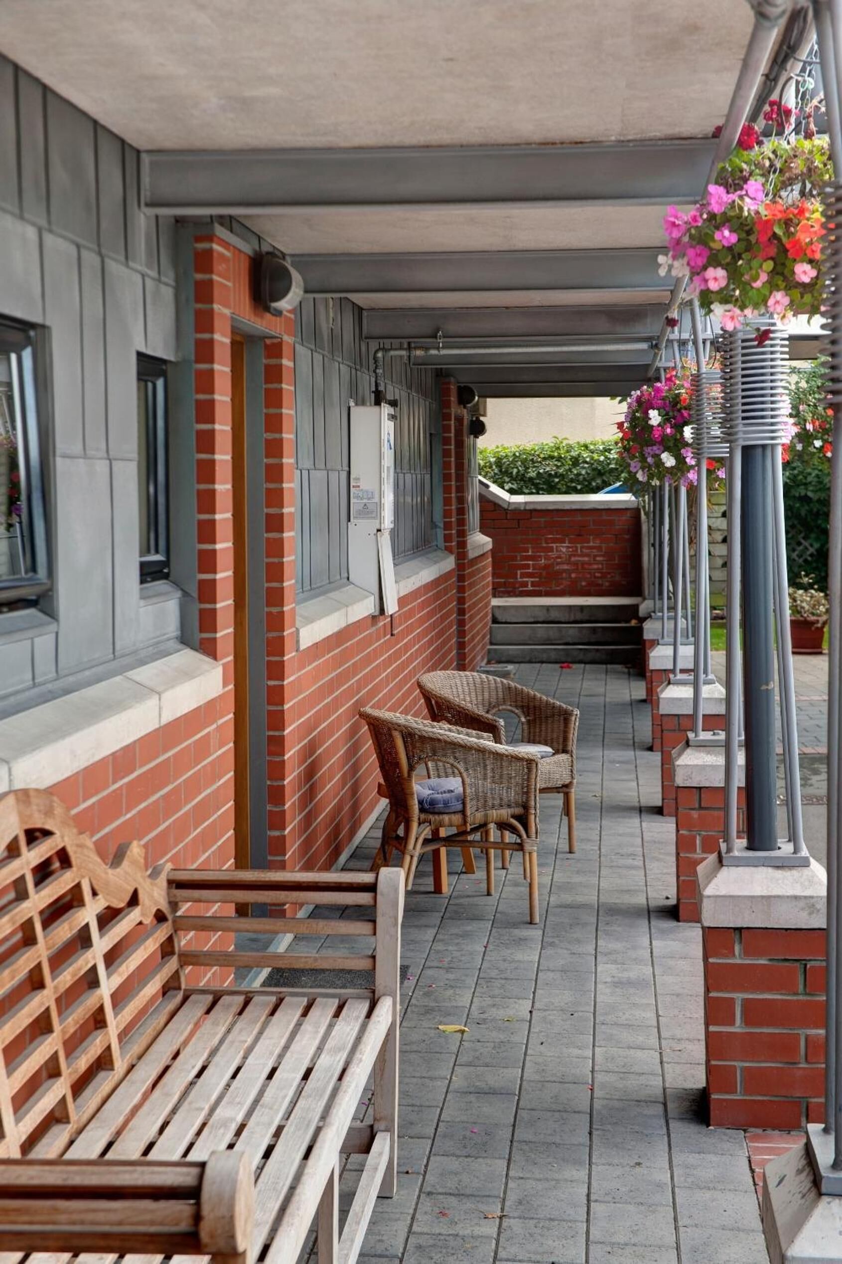 A covered outdoor seating area with wooden benches and wicker chairs, a brick wall, and hanging flower baskets.
