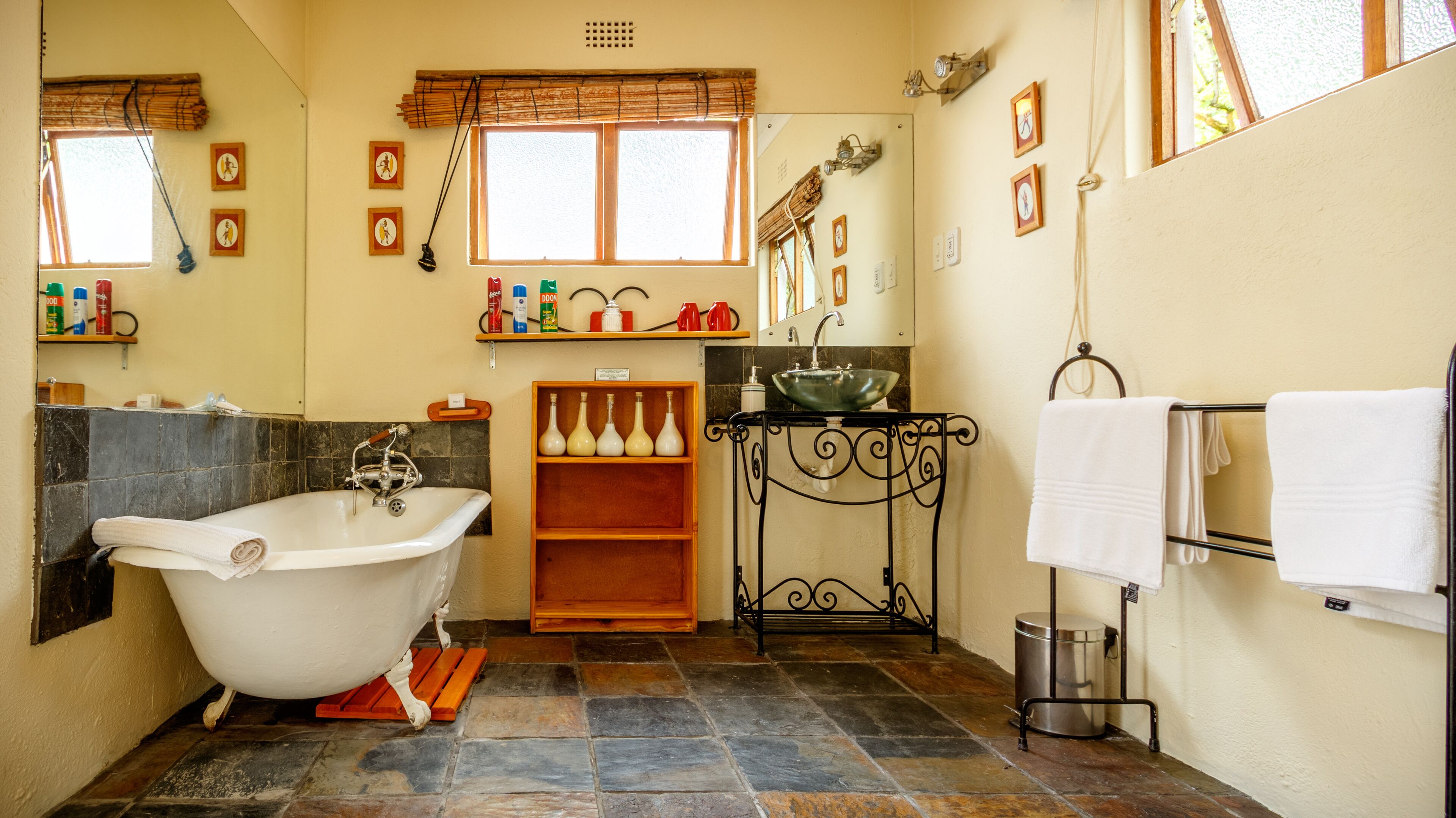 Un baño con una bañera independiente, un lavamanos sobre un soporte de metal negro y un espejo sobre el lavamanos. Hay estantes con artículos de aseo y una ventana con persianas.