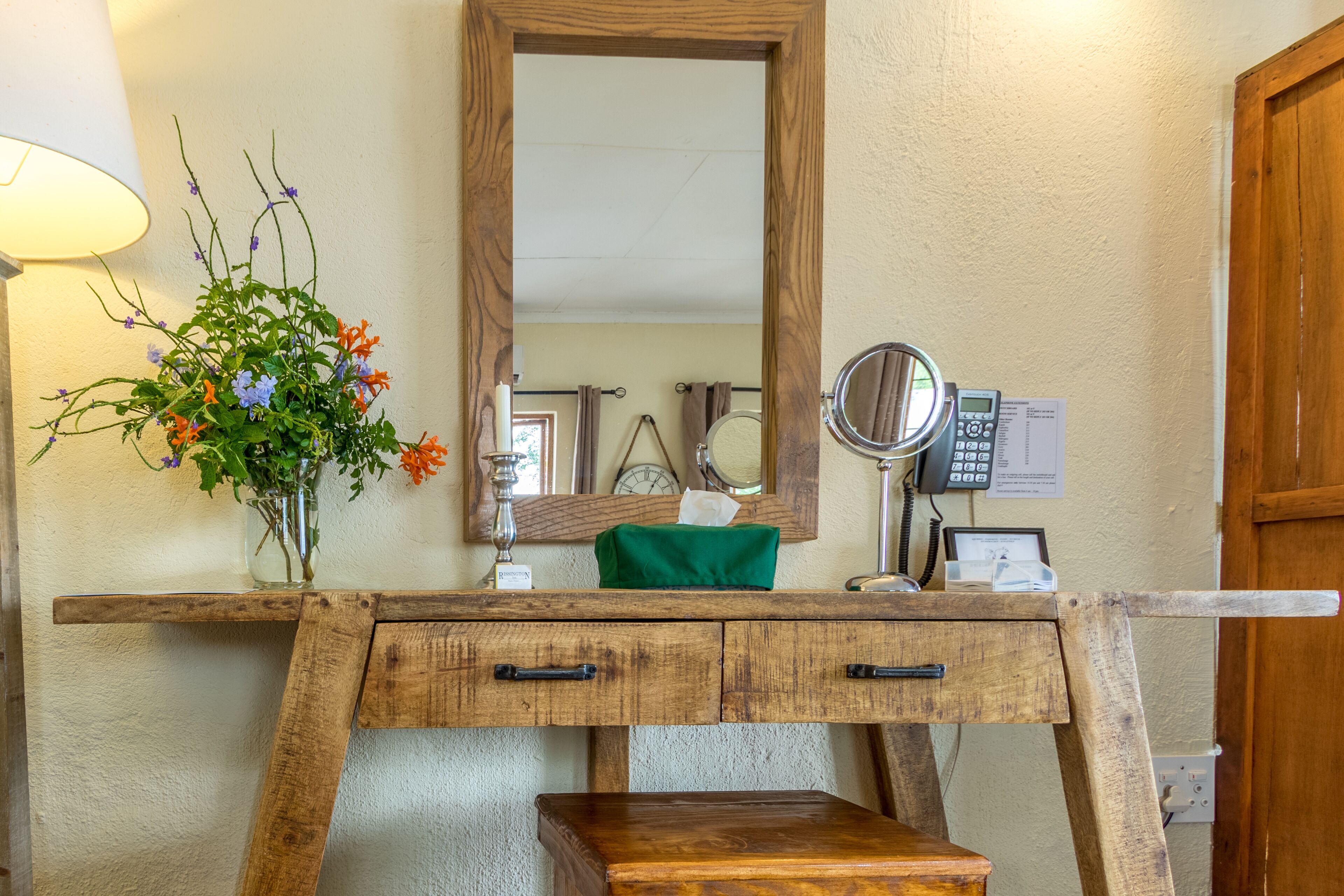A wooden vanity with a mirror, a vase of flowers, and a tissue box.