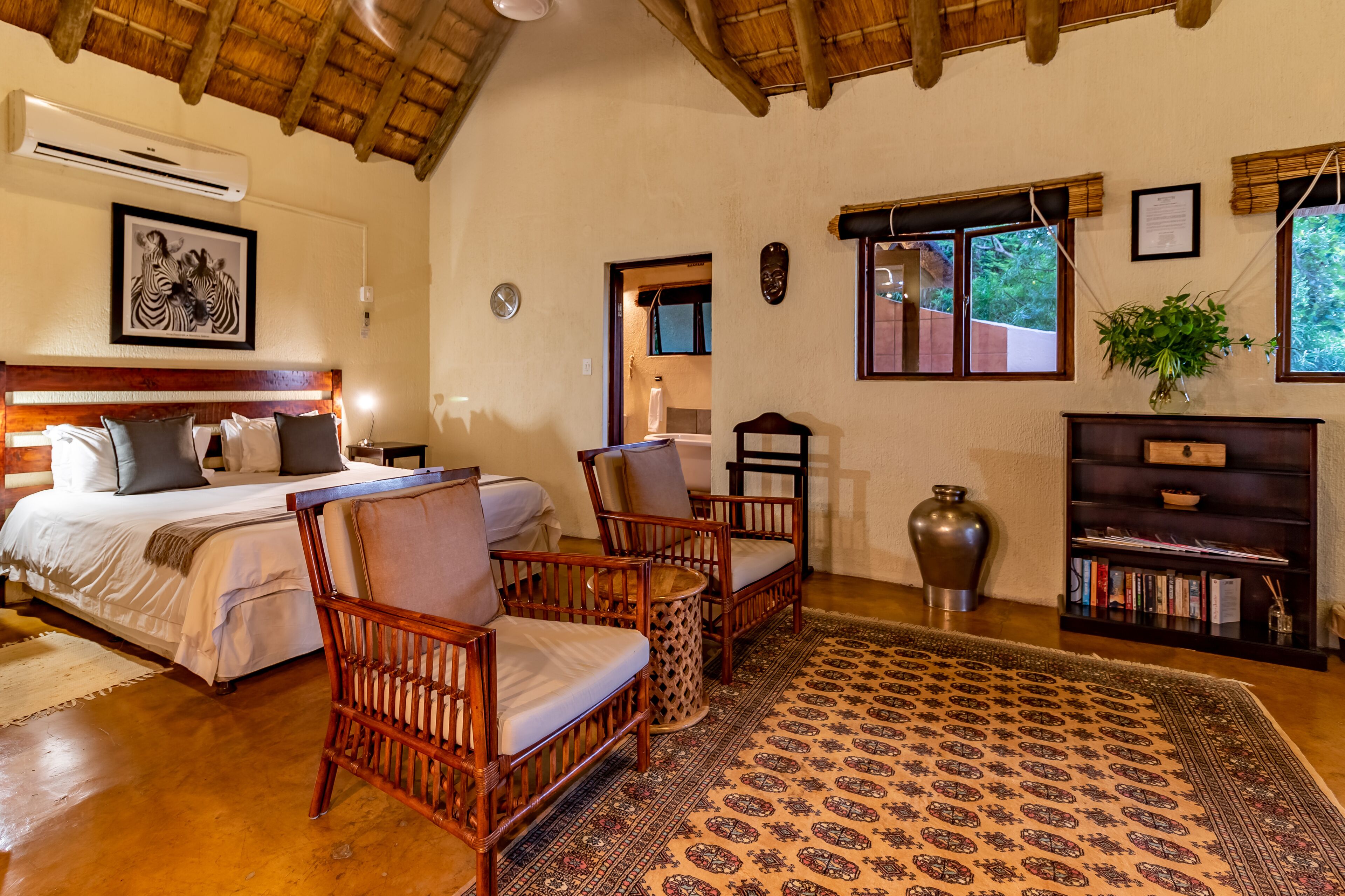 A room with a bed, two wicker chairs, a bookshelf, a large vase, and a patterned rug.