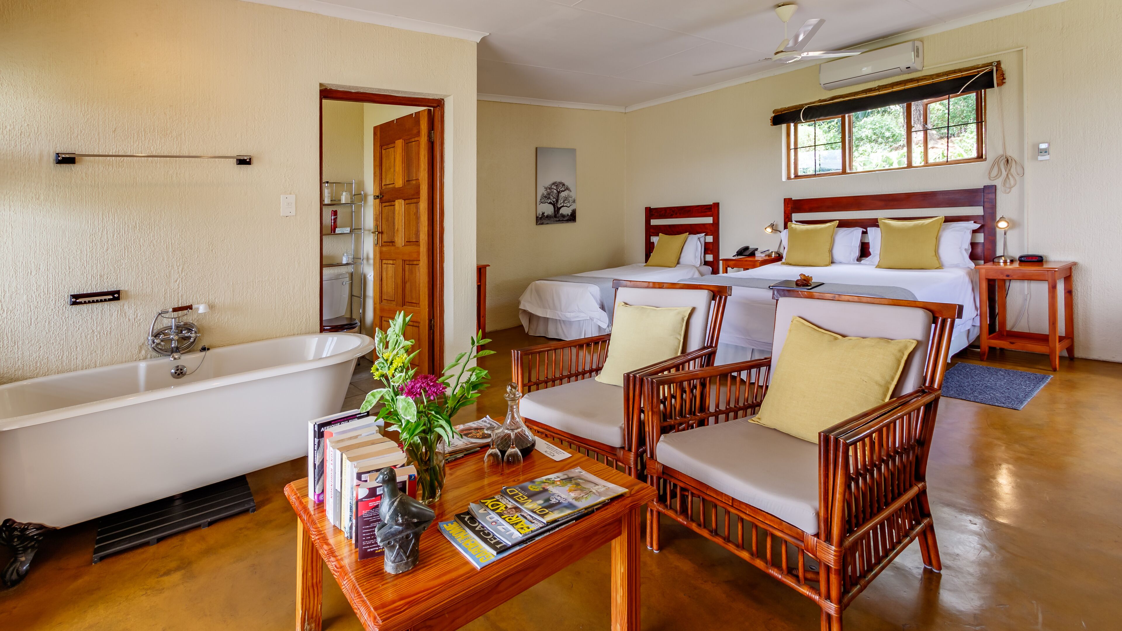 A hotel room with a bathtub, two single beds, a sitting area with wooden furniture, and a large window.