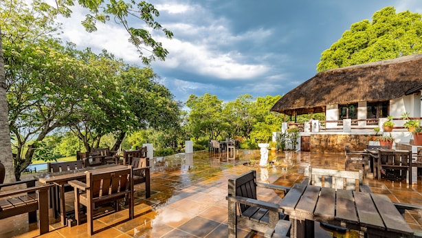 An outdoor dining area with wooden tables and chairs, a thatched-roof building, and lush greenery.
