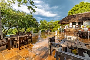 An outdoor dining area with wooden tables and chairs, a thatched-roof building, and lush greenery.