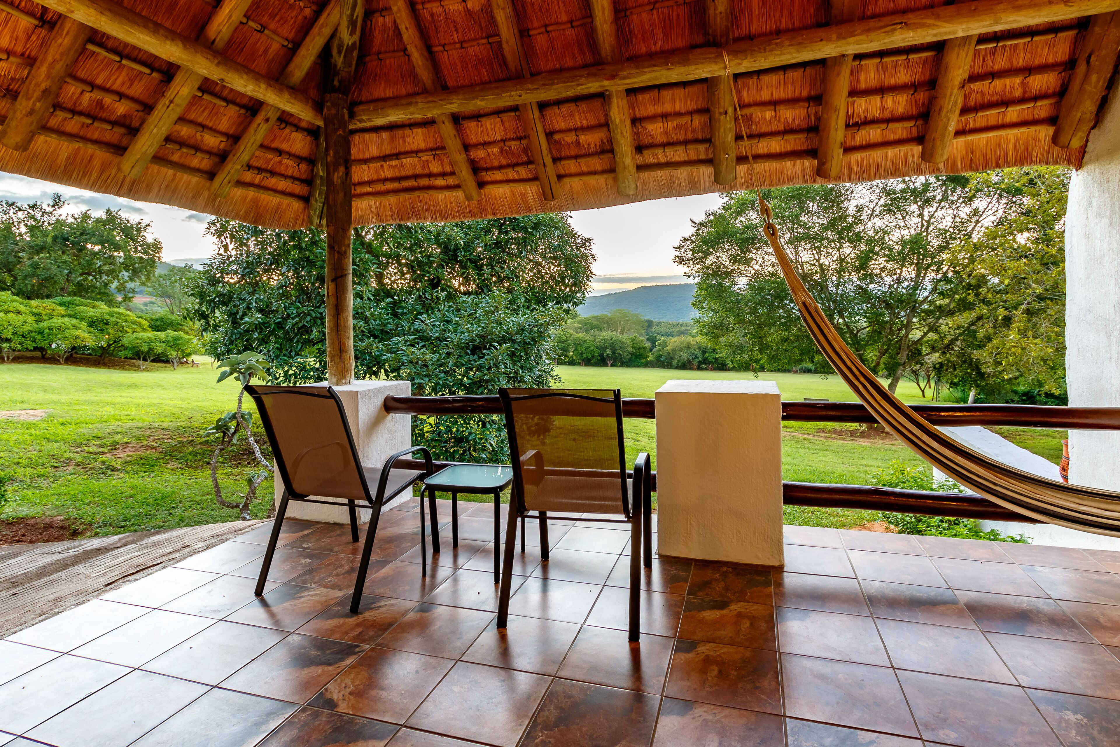 A covered outdoor seating area with a thatched roof, a hammock, and a view of a lush green landscape.