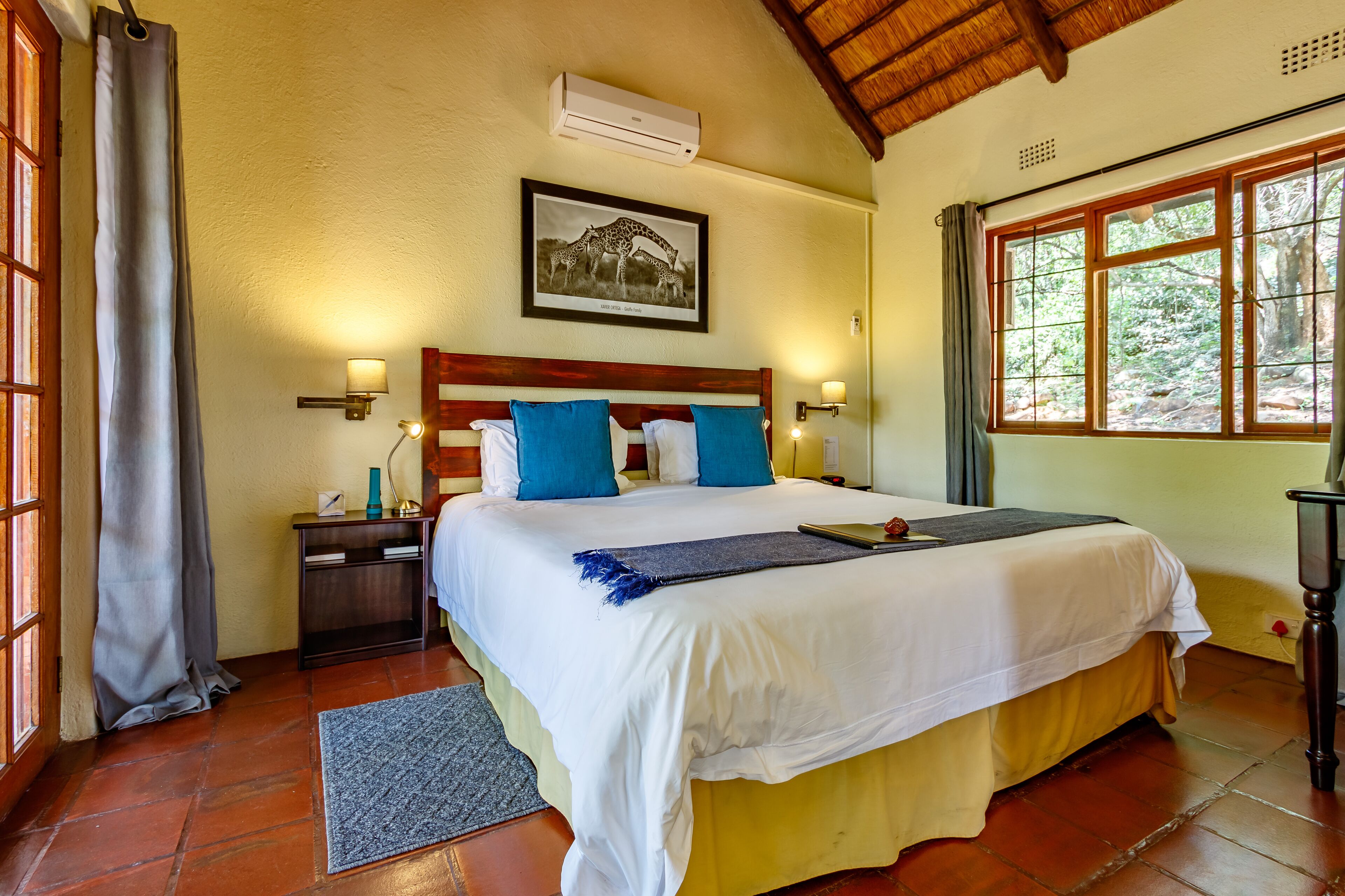 A hotel room with a large bed, a wooden headboard, a framed picture on the wall, and a window with a view of trees.