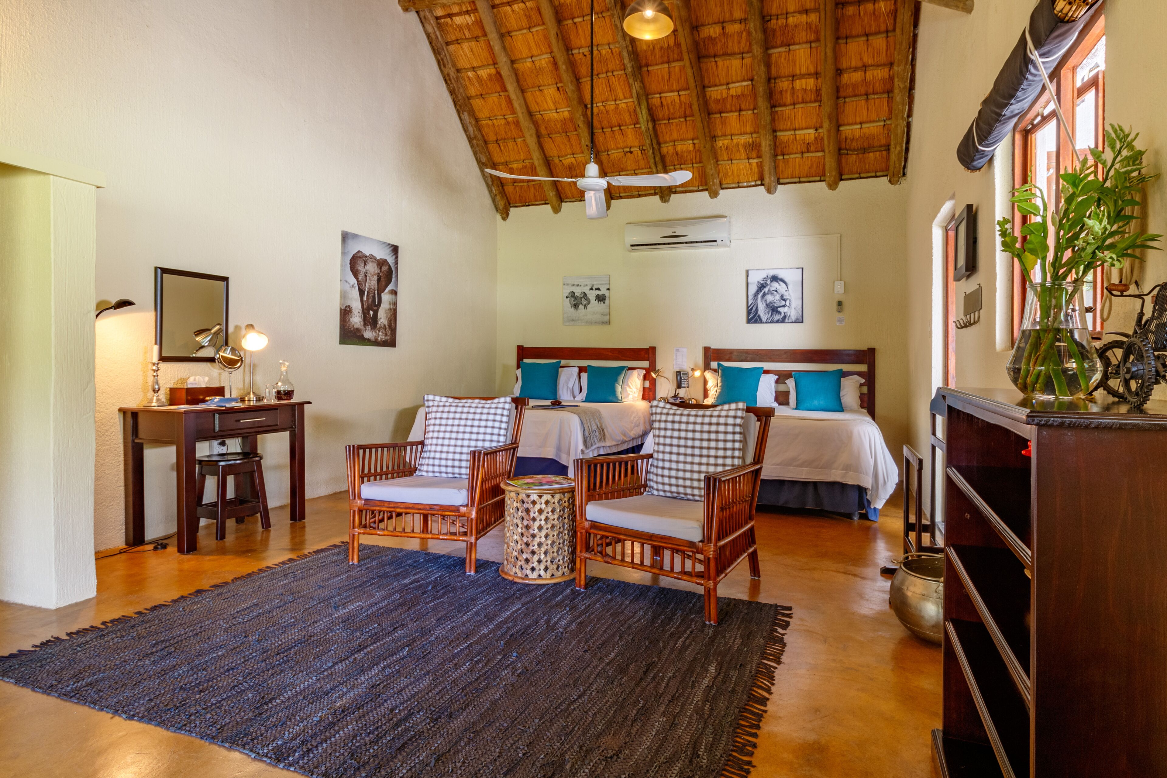 A spacious room with a high ceiling, a bed with a wooden headboard, two wicker chairs, a small round table, and a large potted plant.