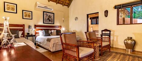 A hotel room with a bed, two wooden chairs, a table with glasses, a large vase, and a window with a view of greenery.