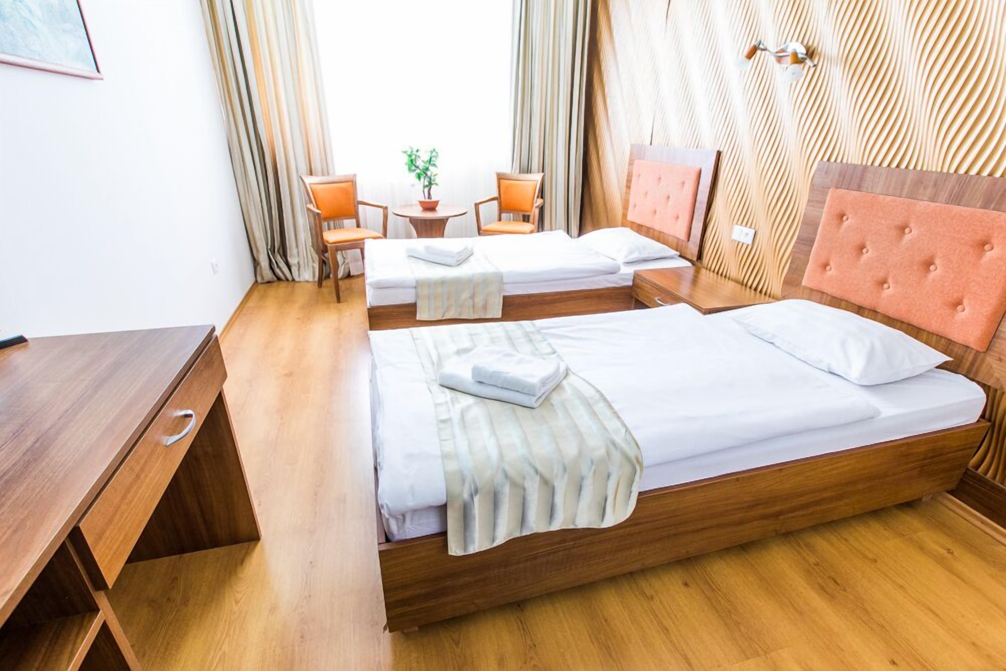 A hotel room with two single beds, a wooden desk, a small table with chairs, and a potted plant.