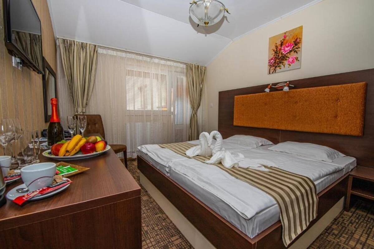 A hotel room with a bed, a desk with breakfast items, a painting, and a window with curtains.