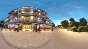 A multi-story hotel with a curved facade, outdoor seating area, and a well-lit entrance. - Hotel Sun (Senec)