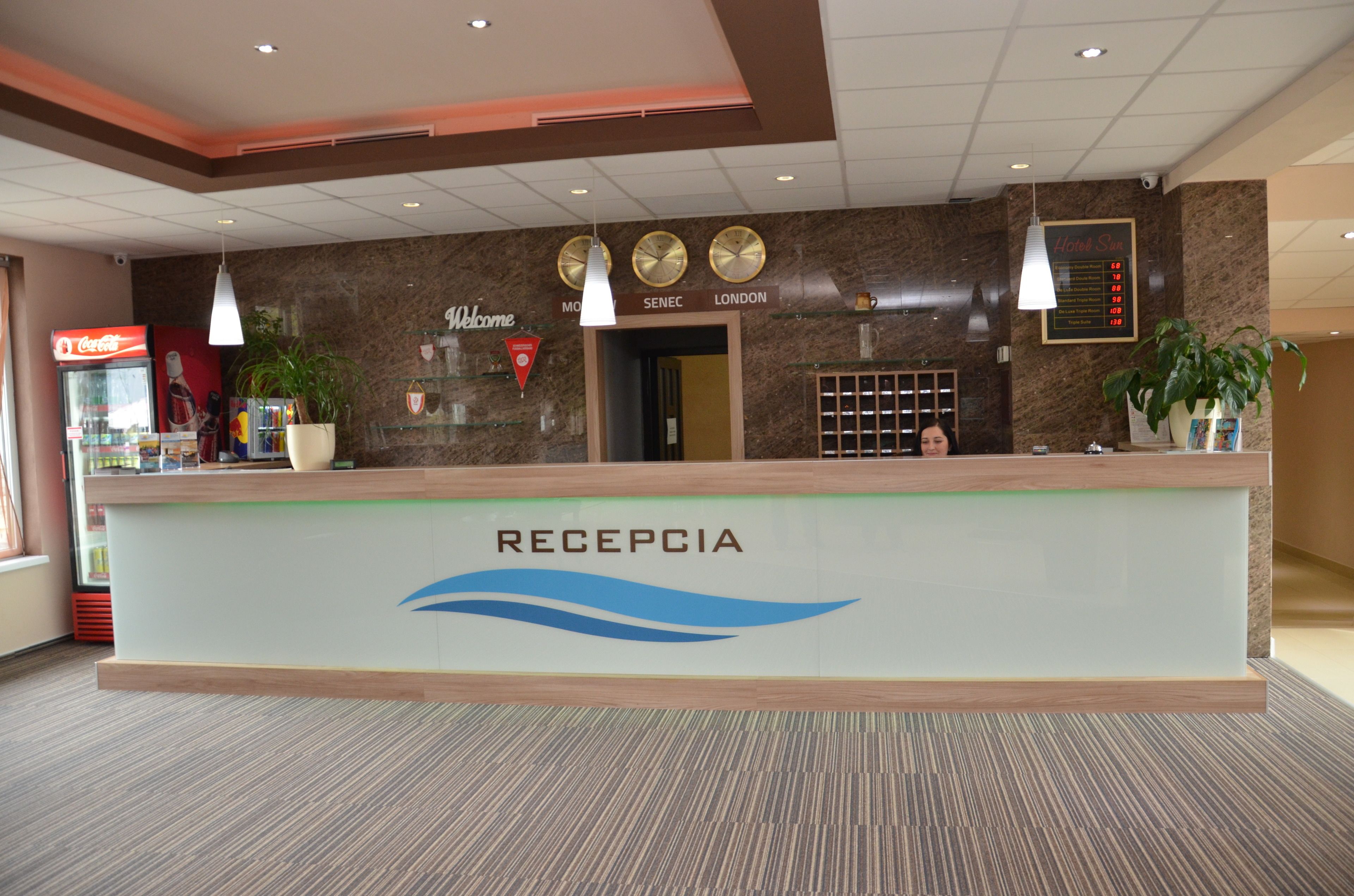 A hotel reception area with a reception desk labeled "RECEPCIA," clocks, a Coca-Cola refrigerator, and a "Welcome" sign.