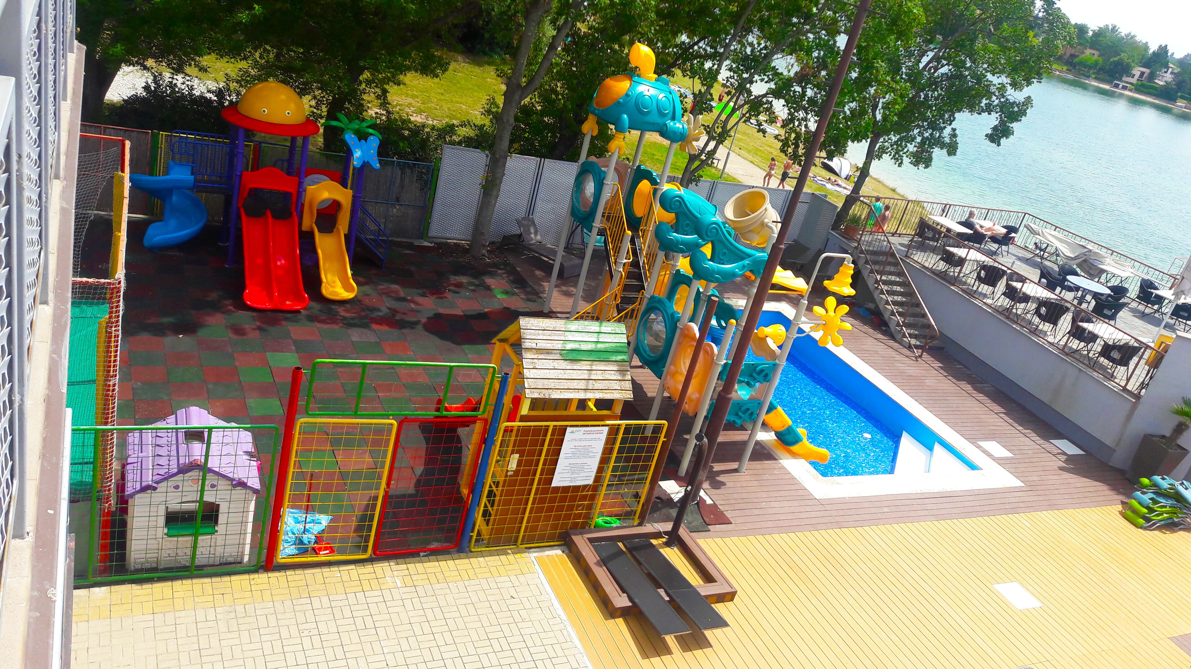 Zona infantil con toboganes y estructuras de escalada, piscina y mobiliario exterior junto al agua.