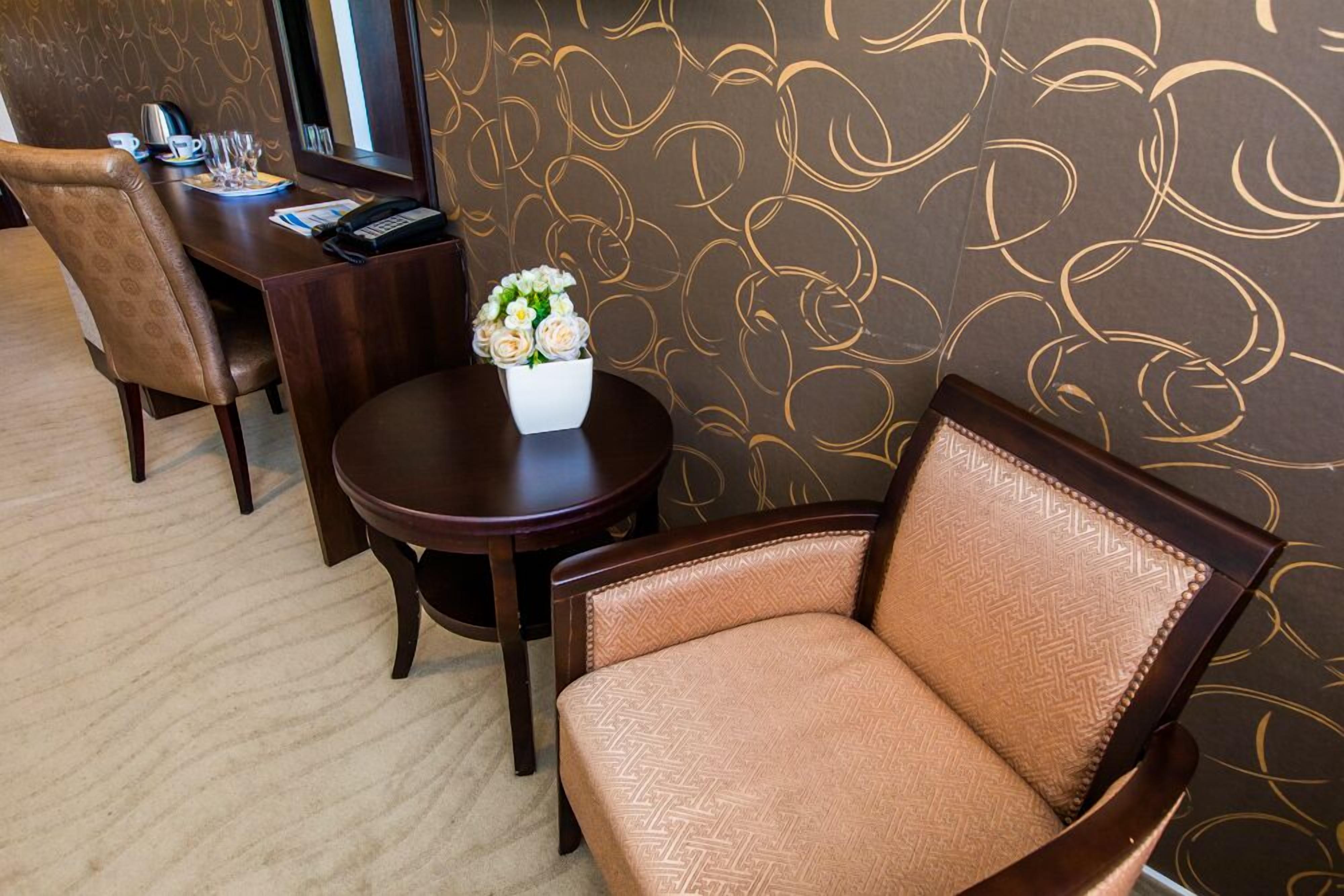 Quarto de hotel com parede em tom de castanho com padrão, uma mesa redonda com um vaso de flores, uma poltrona de couro marrom e uma escrivaninha de madeira com telefone e papéis.
