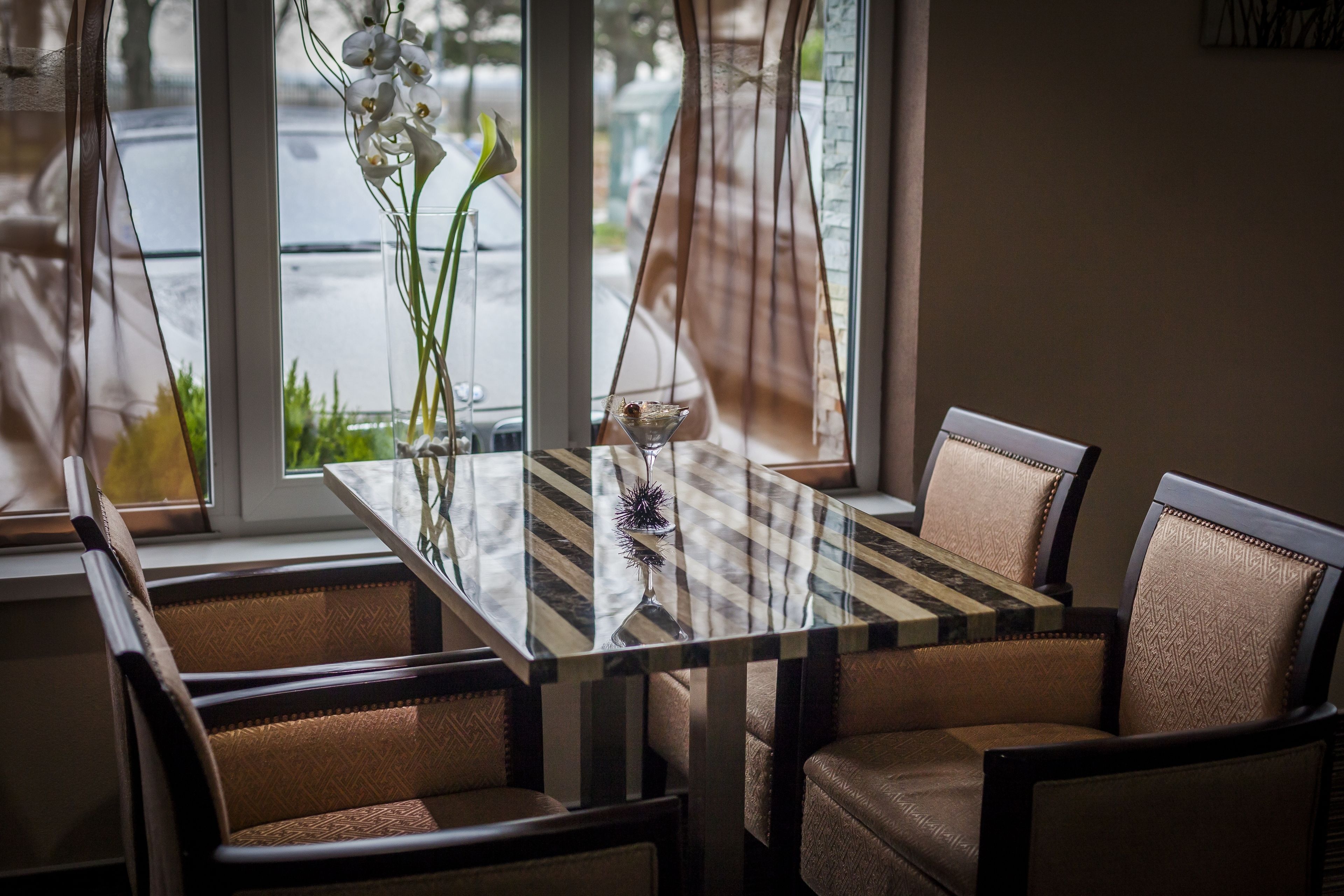 A dining area with a glass table and cushioned chairs, a vase with flowers, and a view of the street through large windows.