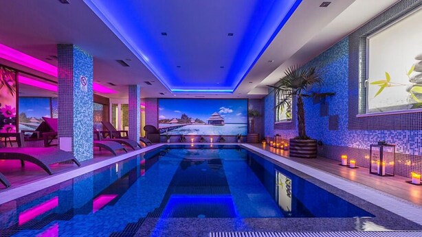 A modern indoor swimming pool with a view of a tropical landscape, surrounded by lounge chairs and ambient lighting. - Hotel Sun (Senec)