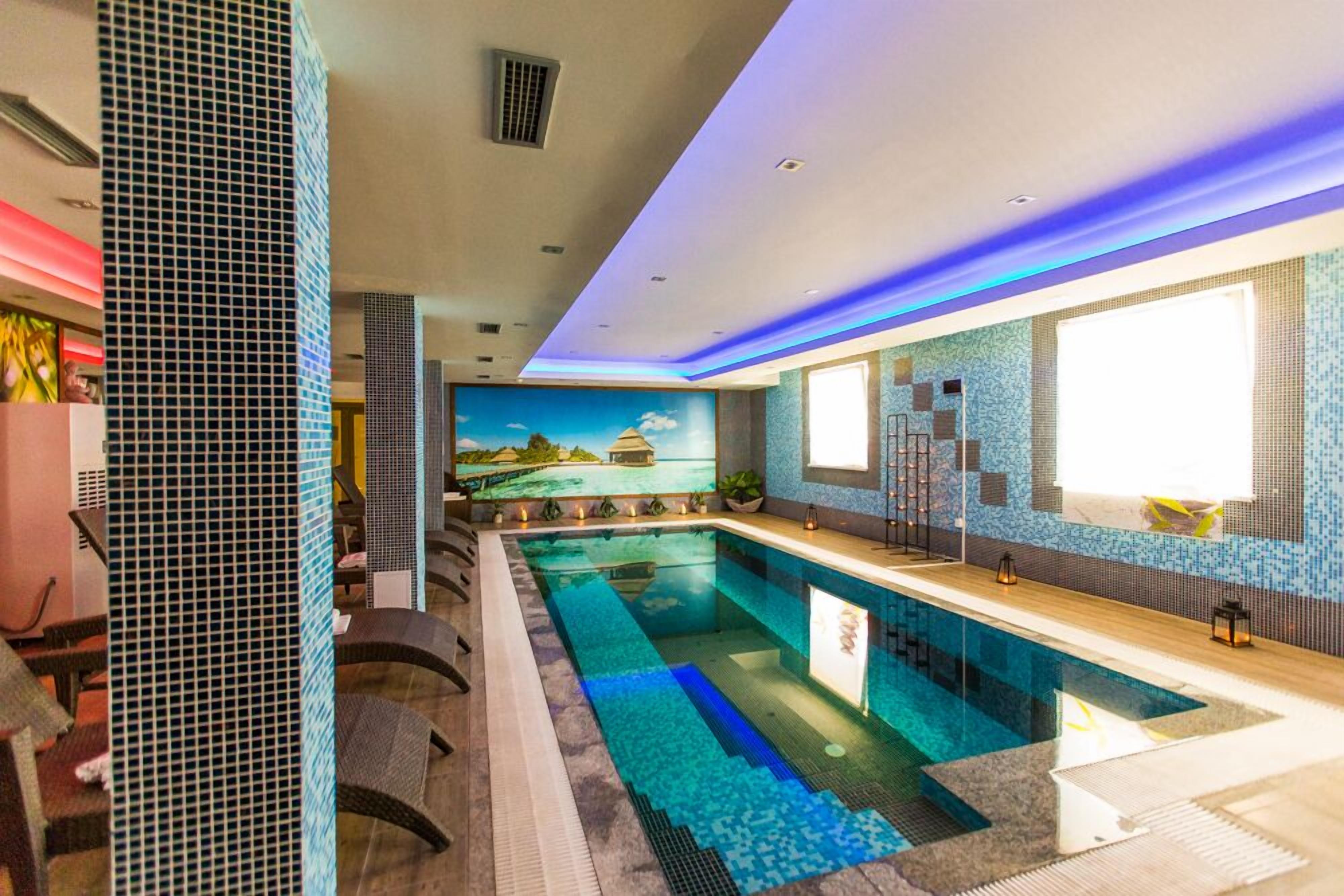 A modern indoor swimming pool with a long, narrow design, surrounded by a tiled wall with a mosaic pattern.