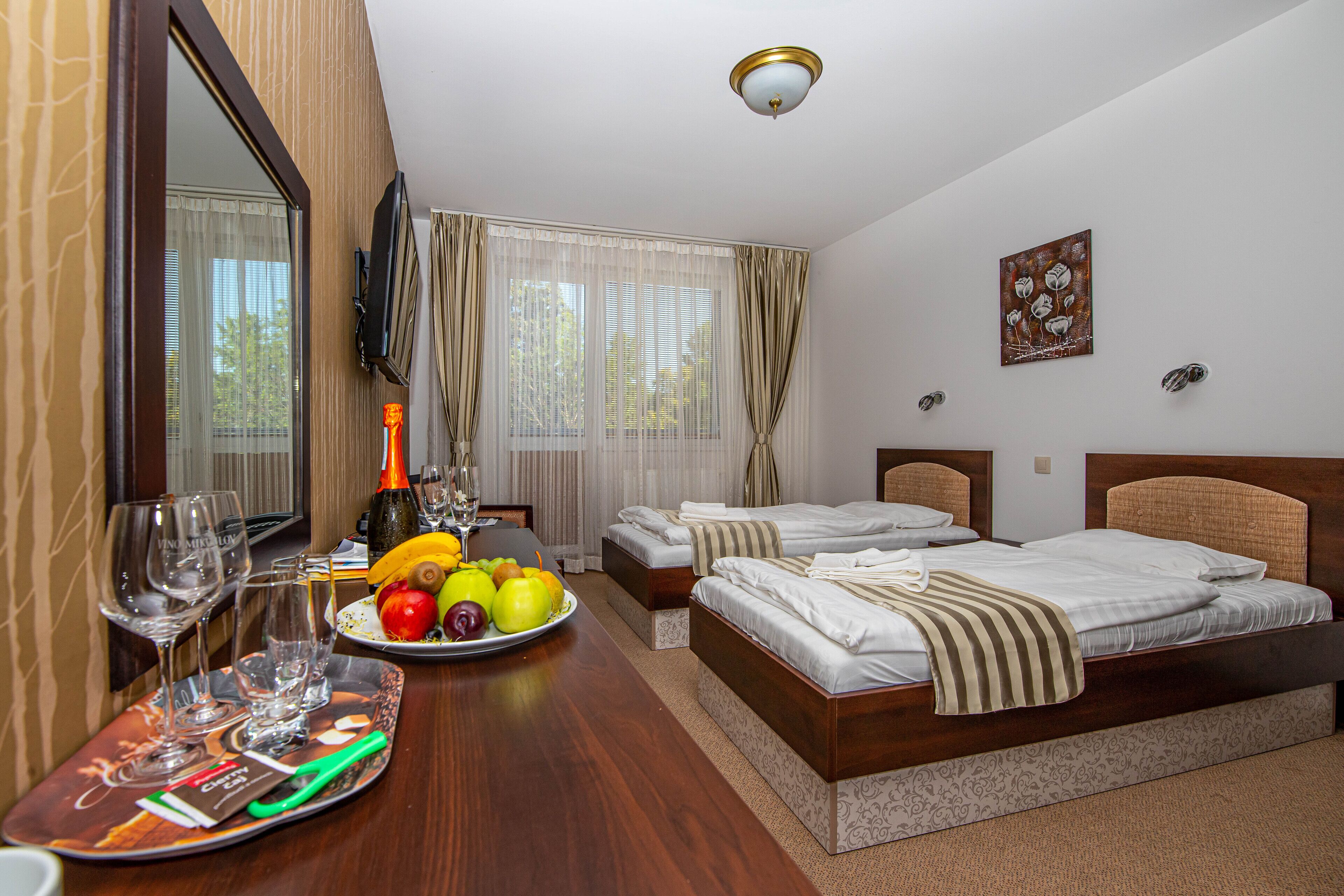 Habitación de hotel con dos camas, una mesa de comedor con frutas y vasos, y un televisor montado en la pared.