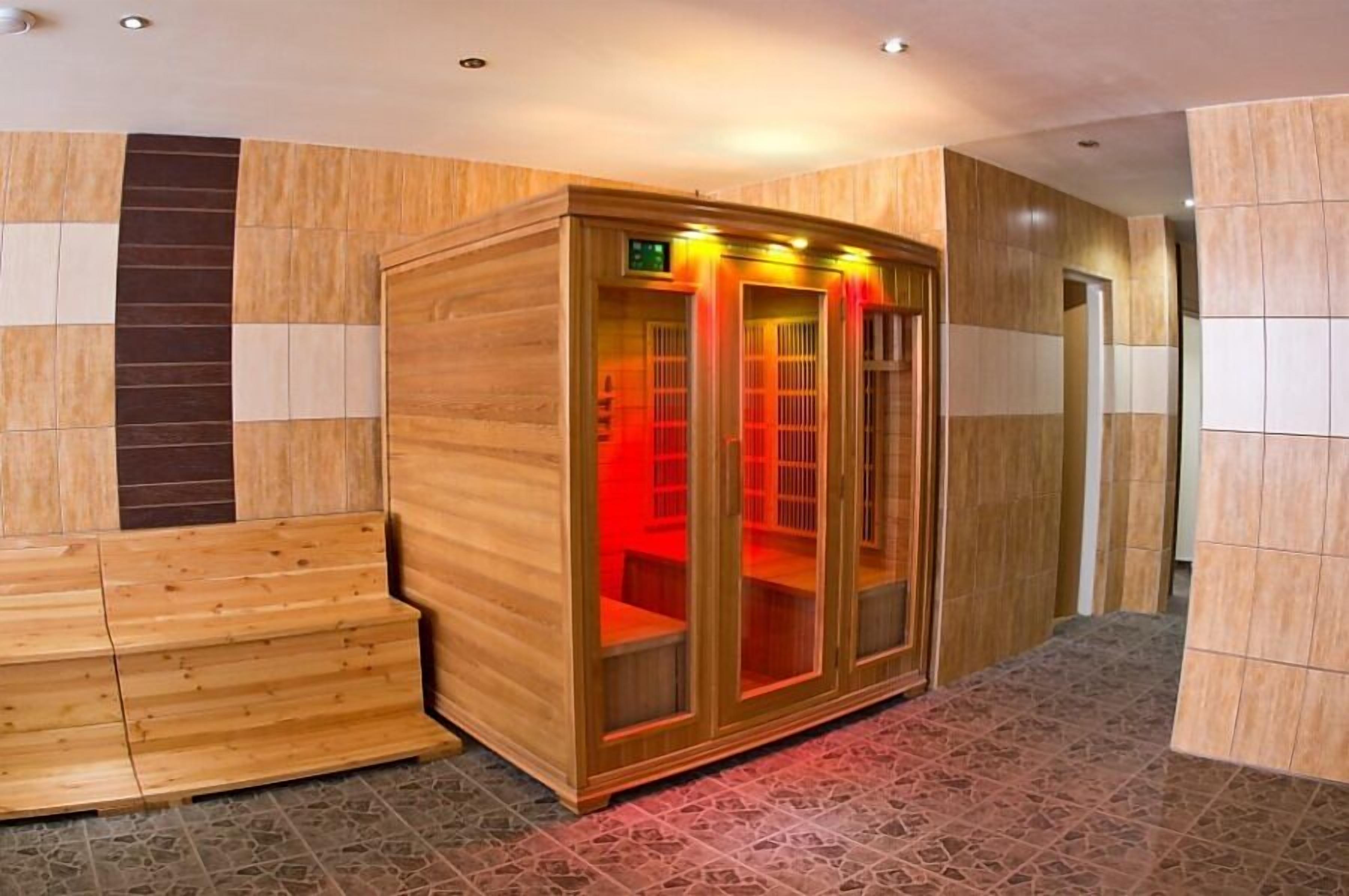 Una sauna con bancos de madera y un recinto de vidrio, ubicada en un pasillo con paredes y suelos de baldosas.