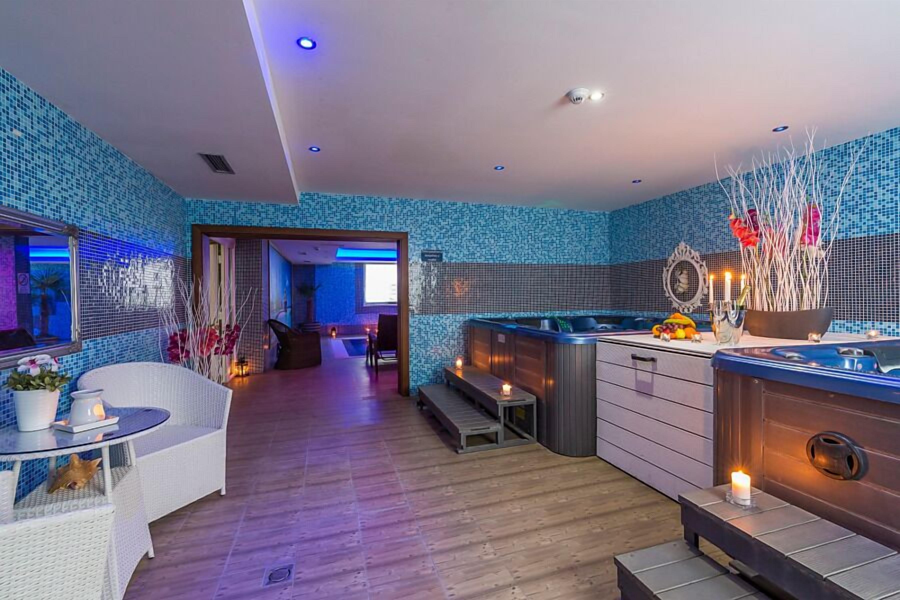 Un espace spa moderne doté d’un plancher en bois, d’un coin salon avec une table et des chaises, d’un jacuzzi et d’un mur décoratif recouvert de carreaux bleus.