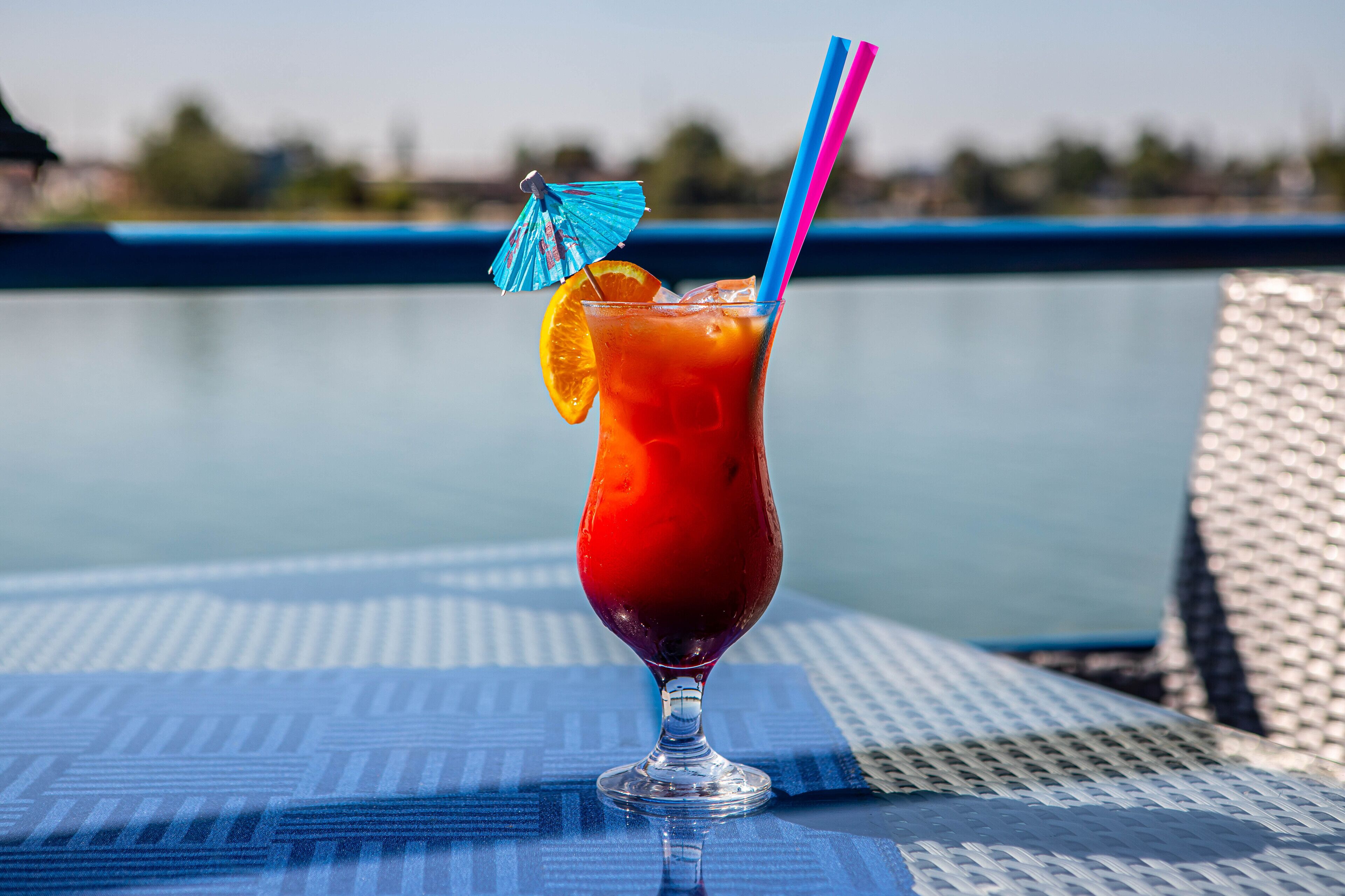 Un vaso de té helado con una rodaja de naranja y un paraguas azul sobre una mesa junto a una piscina.