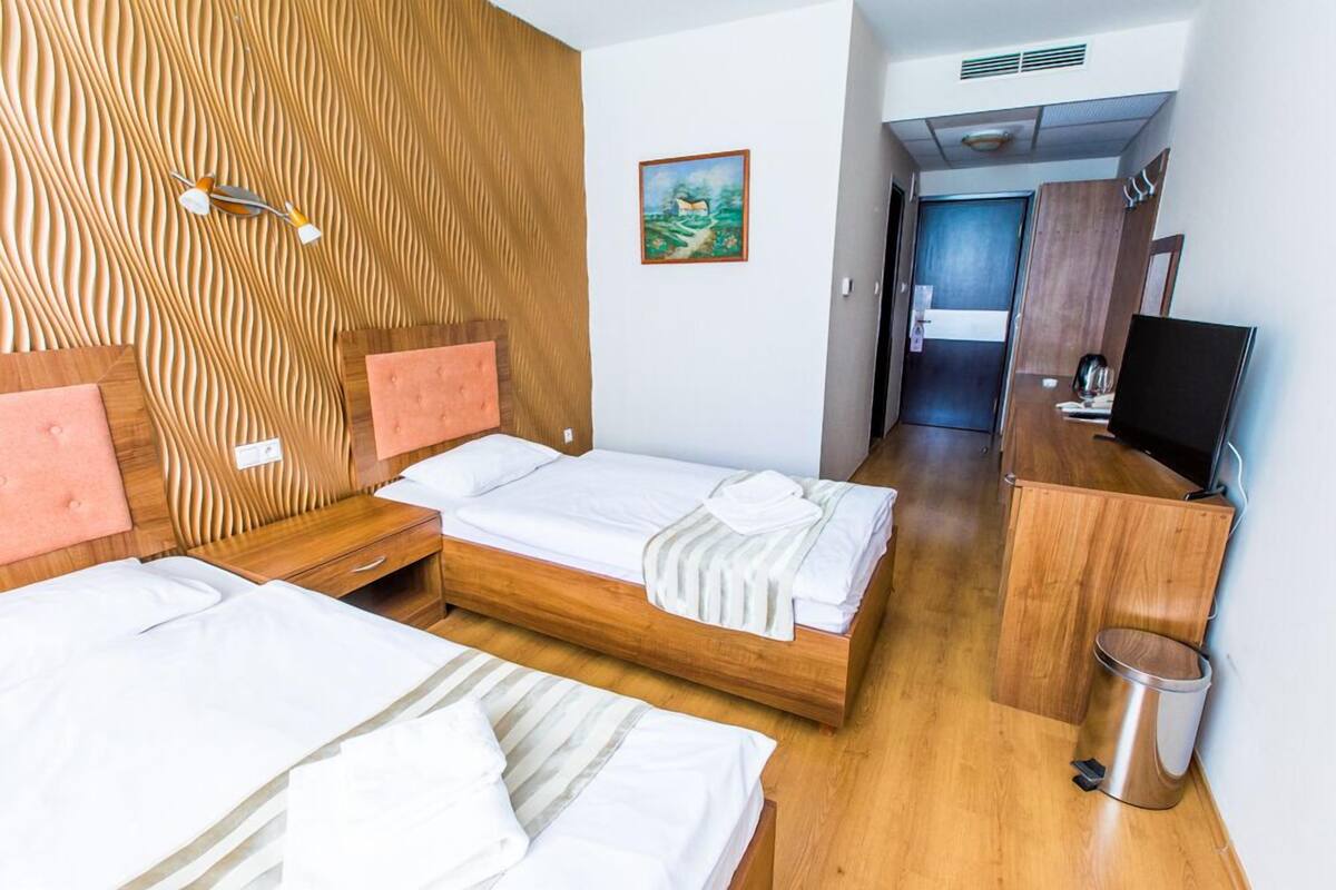 A hotel room with two beds, wooden headboards, a painting on the wall, a television, and a trash bin.