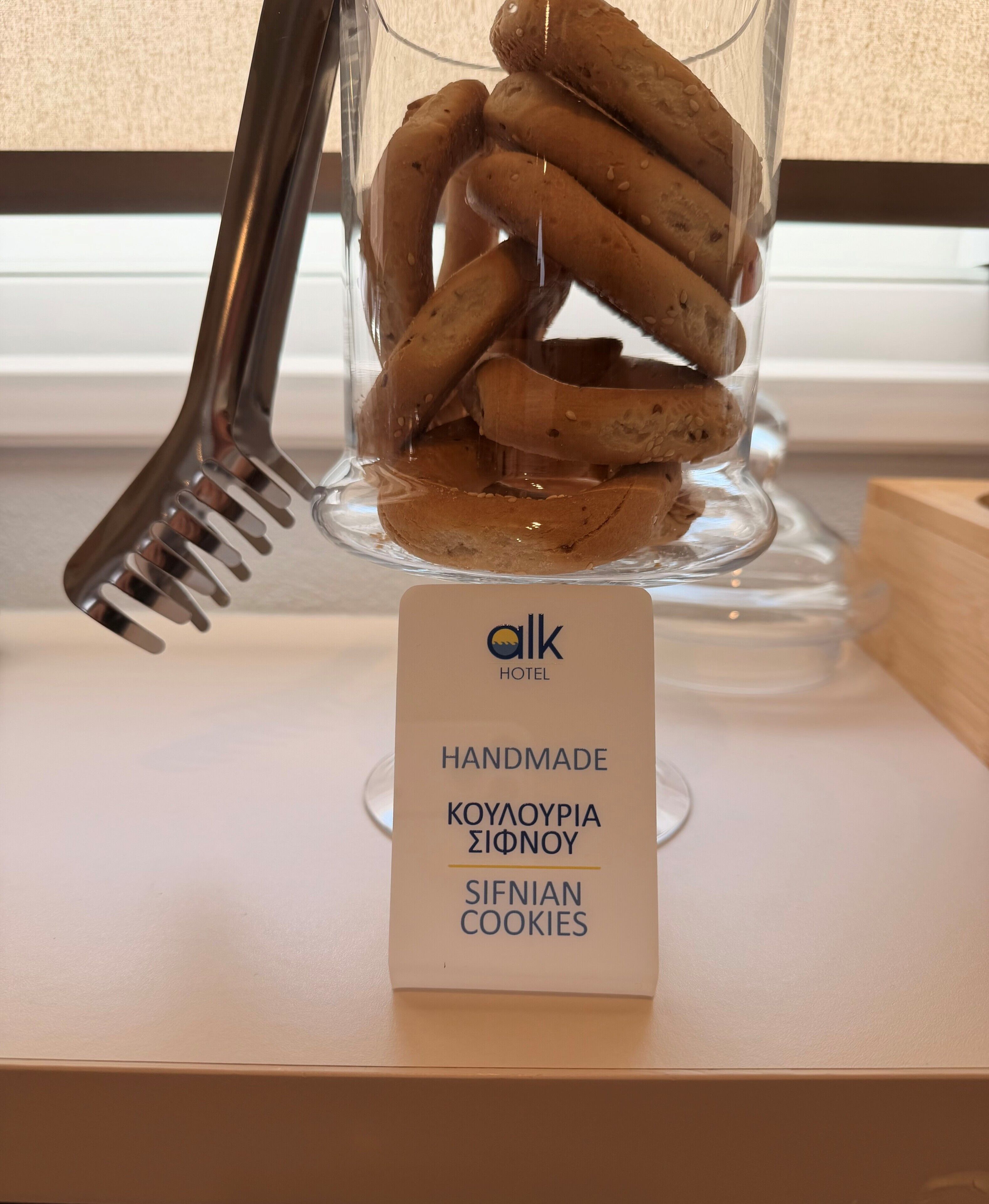 Un tarro de vidrio lleno de galletas, un tenedor de metal y un cartel que indica que son galletas caseras de Sifnos del hotel Alk.