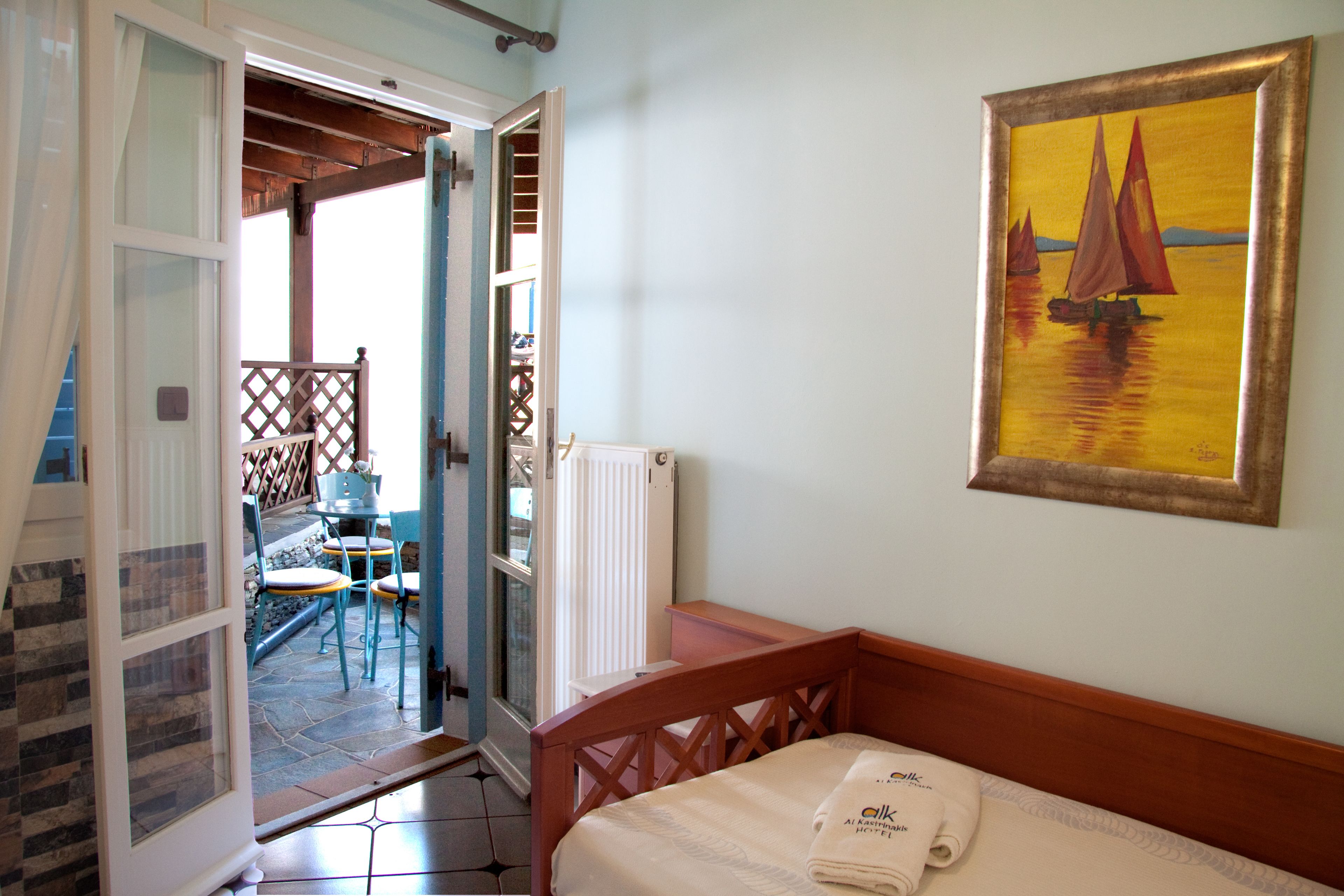 A bedroom with a bed, a painting of sailboats, and a view of an outdoor patio with chairs and a table.