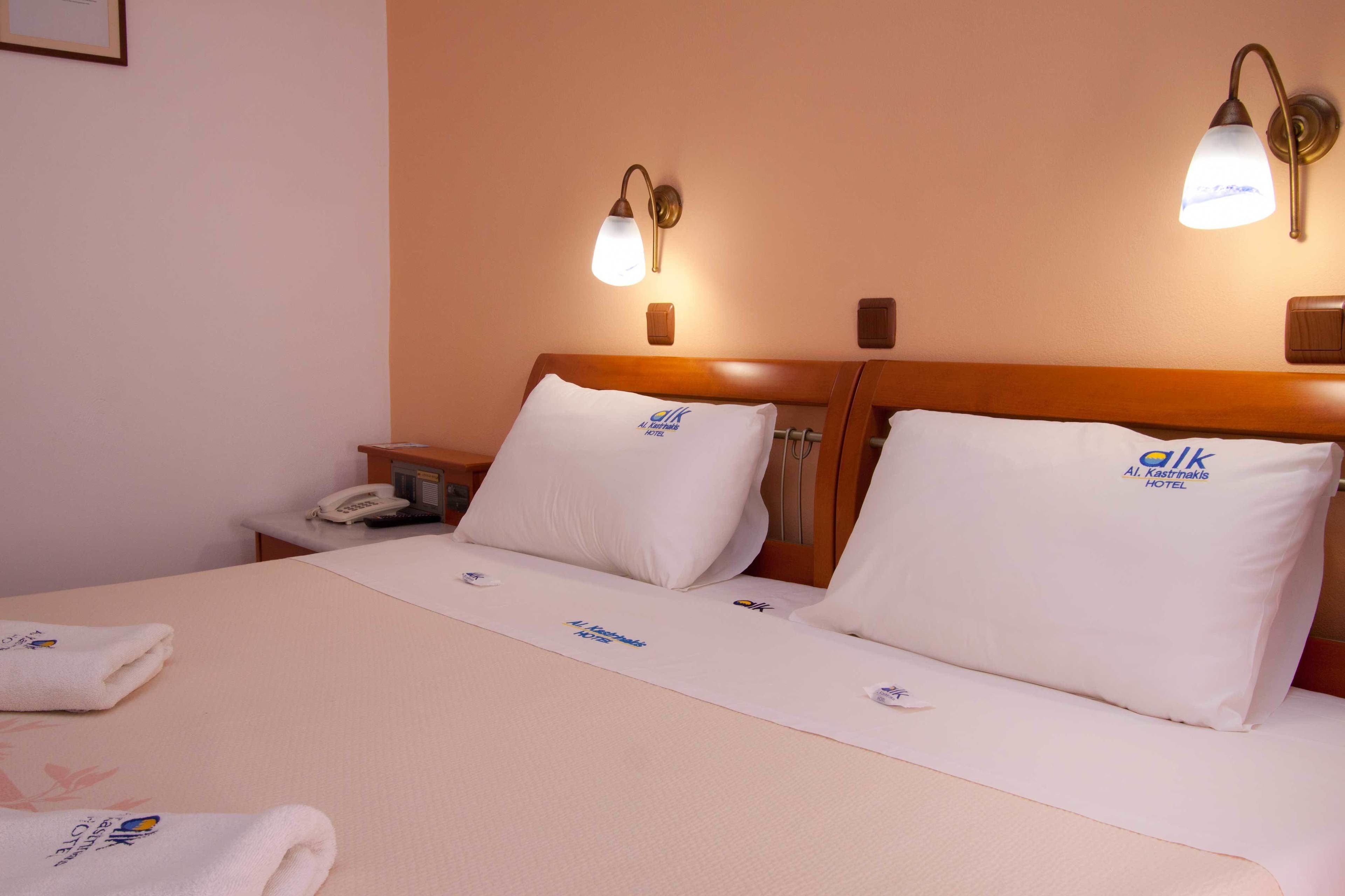 A hotel room with a neatly made bed, two pillows with the hotel logo, a bedside table with a phone, and wall-mounted lamps.
