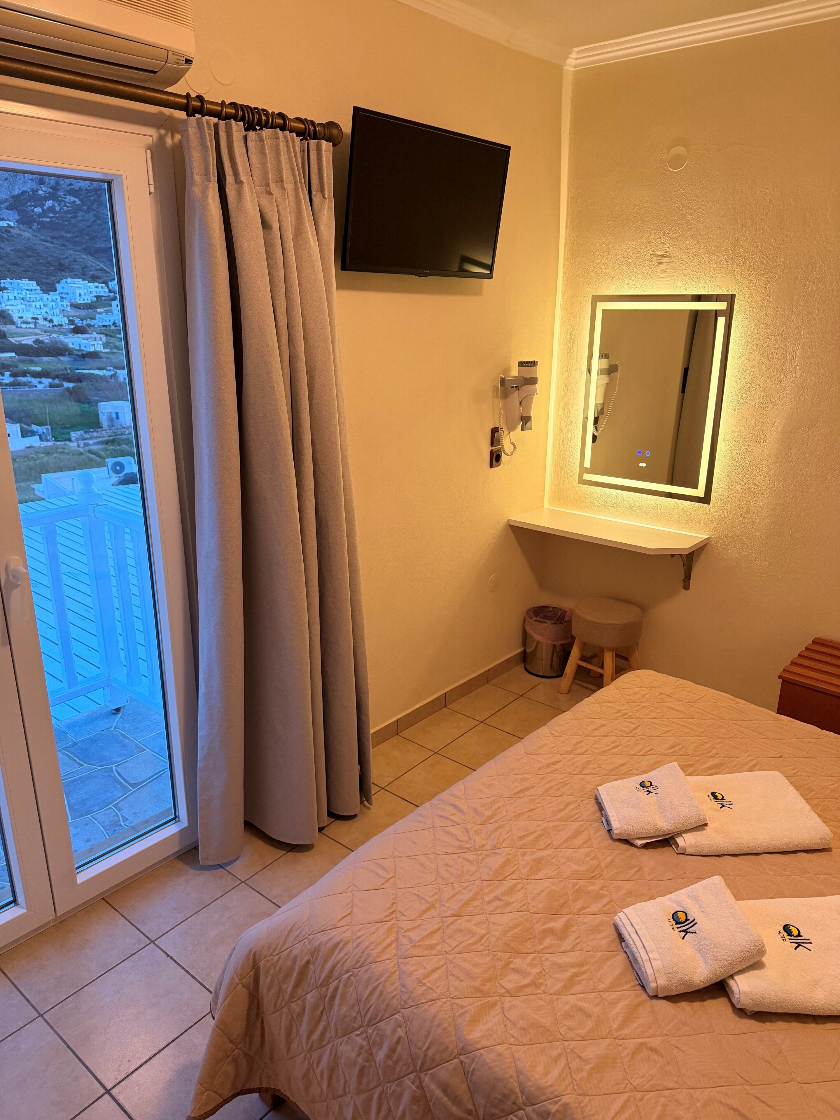 A hotel room with a bed, a TV, a mirror, a window with curtains, and a view of buildings outside.