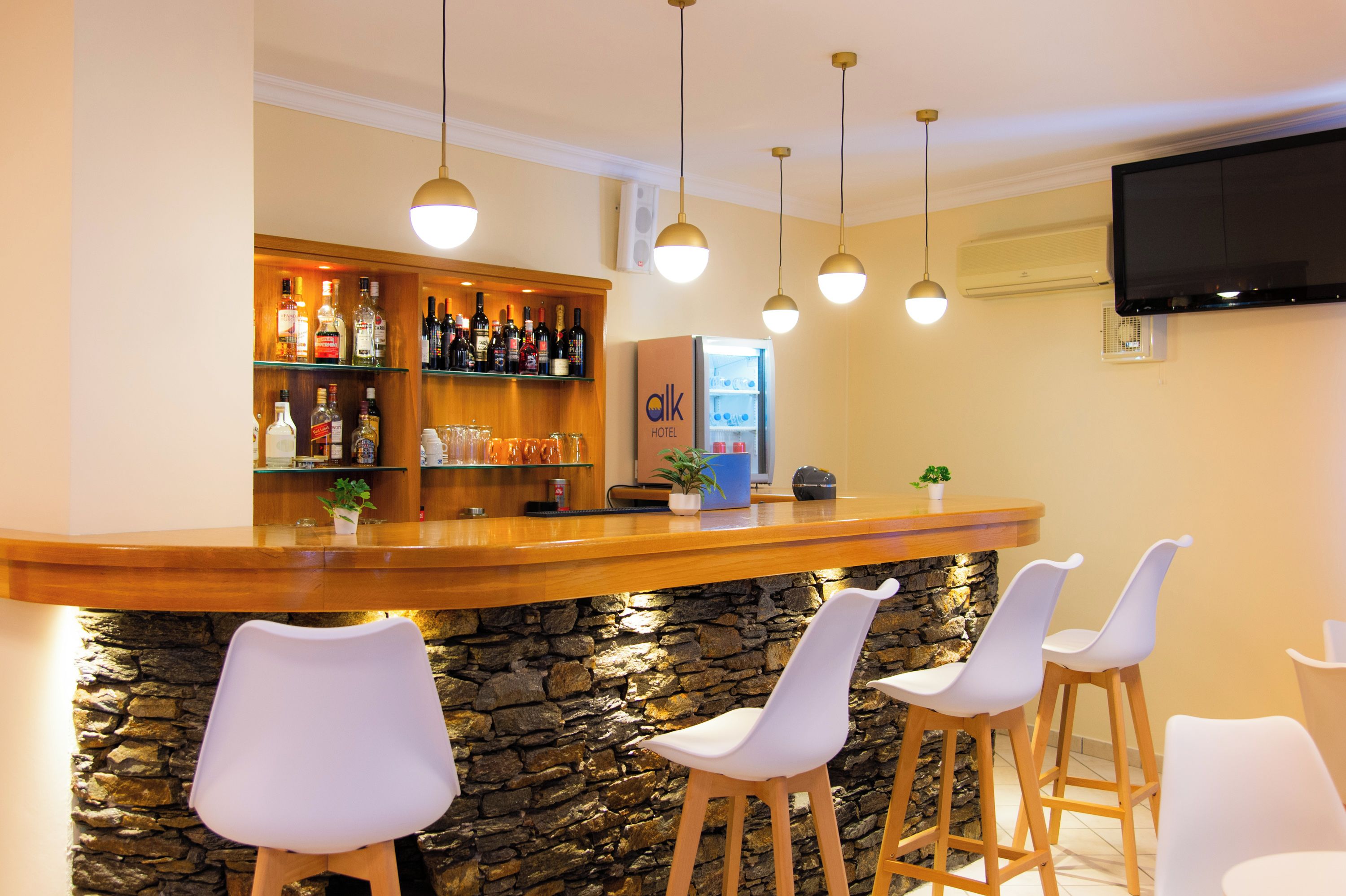 A bar area with a stone wall, wooden counter, and white bar stools. There are hanging pendant lights and a flat-screen TV mounted on the wall.